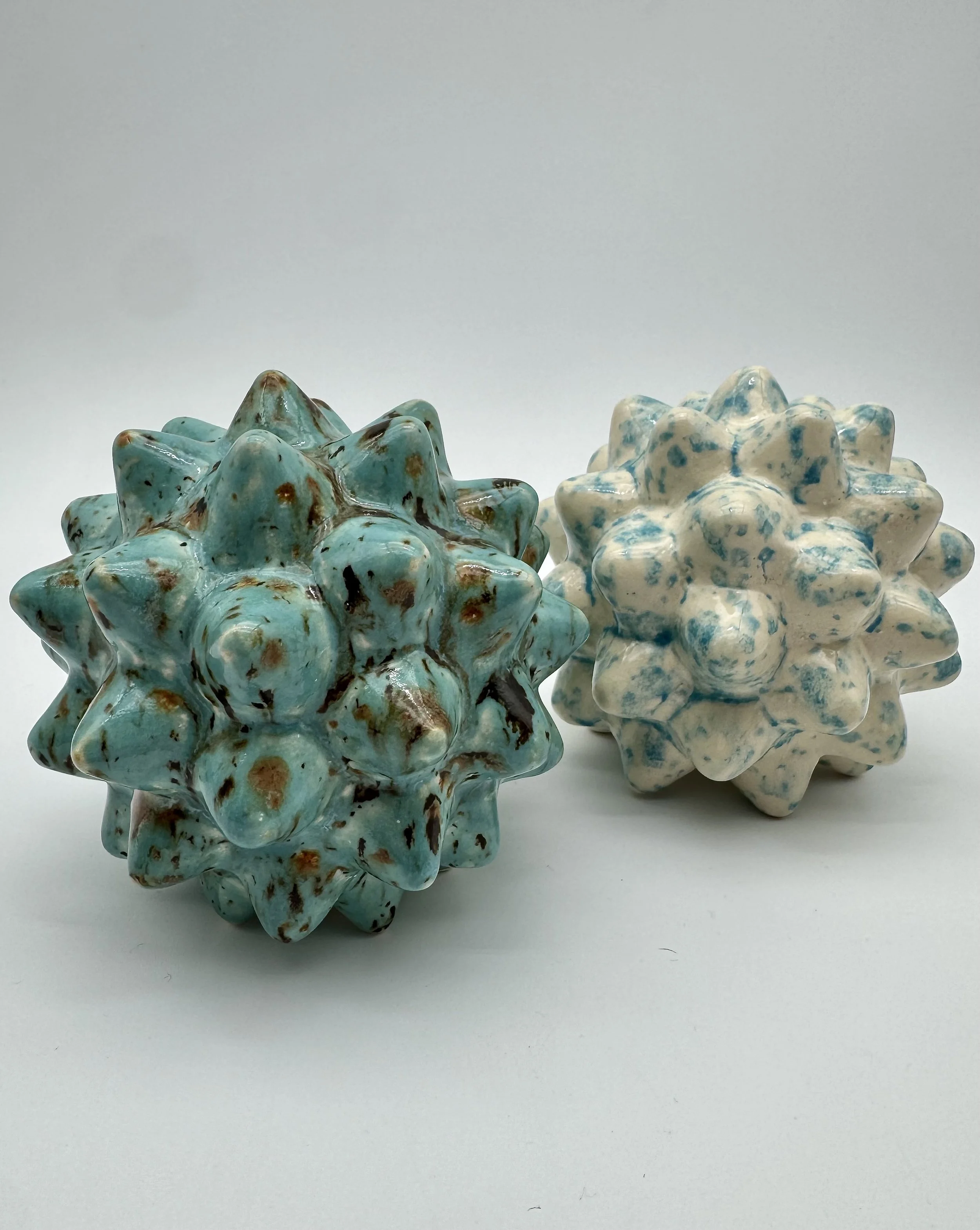 Two ceramic star-shaped sculptures with a glossy glaze, one with turquoise and brown spots, the other with white and blue speckles, placed on a white surface against a plain background.