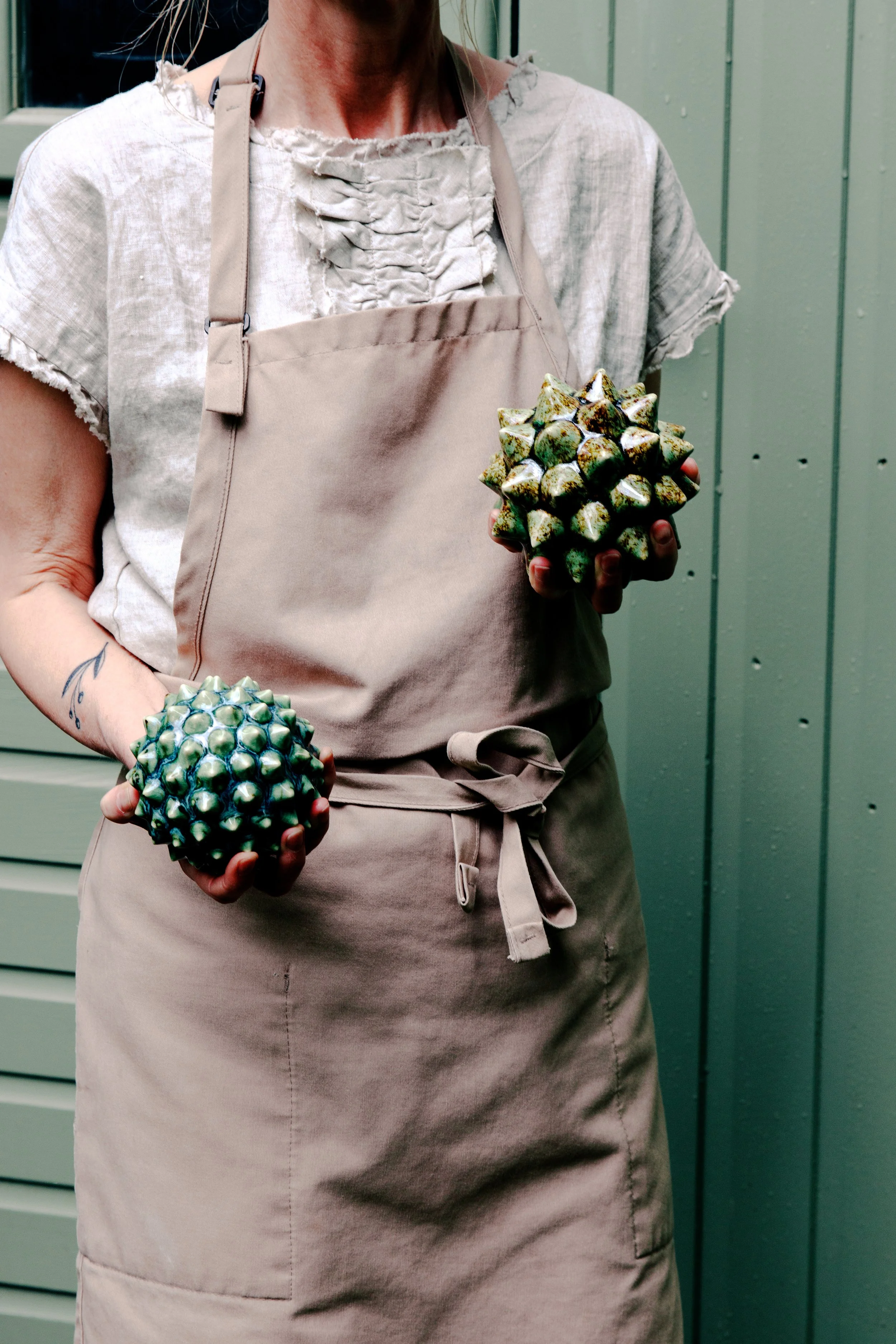 Person wearing a beige apron and holding two ornamental objects resembling large, spiky fruits, standing in front of green wooden panels.