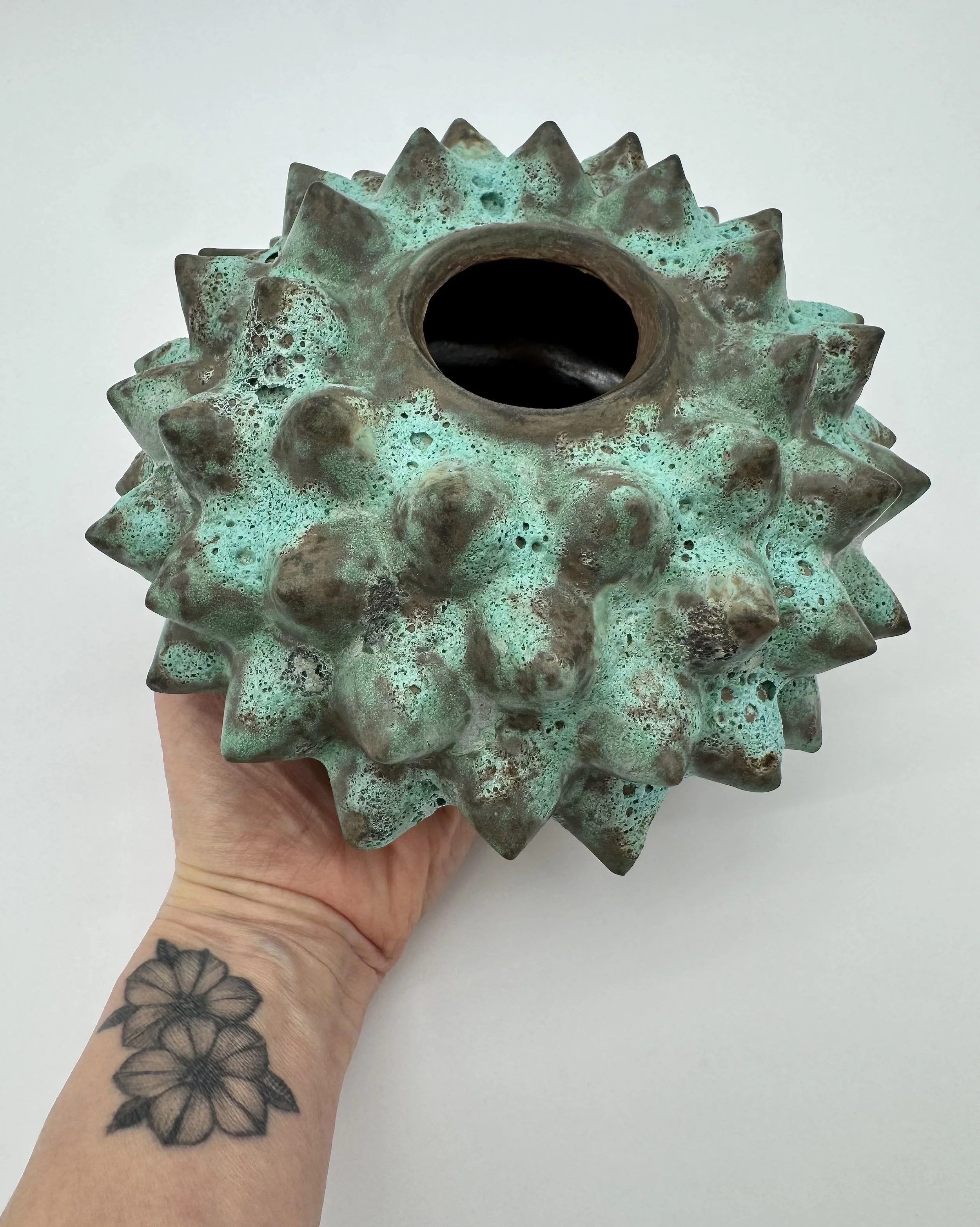 A hand holding a ceramic spiky, spherical object with a large hole in the center. The object has a textured, turquoise and brown surface. The person has a floral tattoo on their forearm.