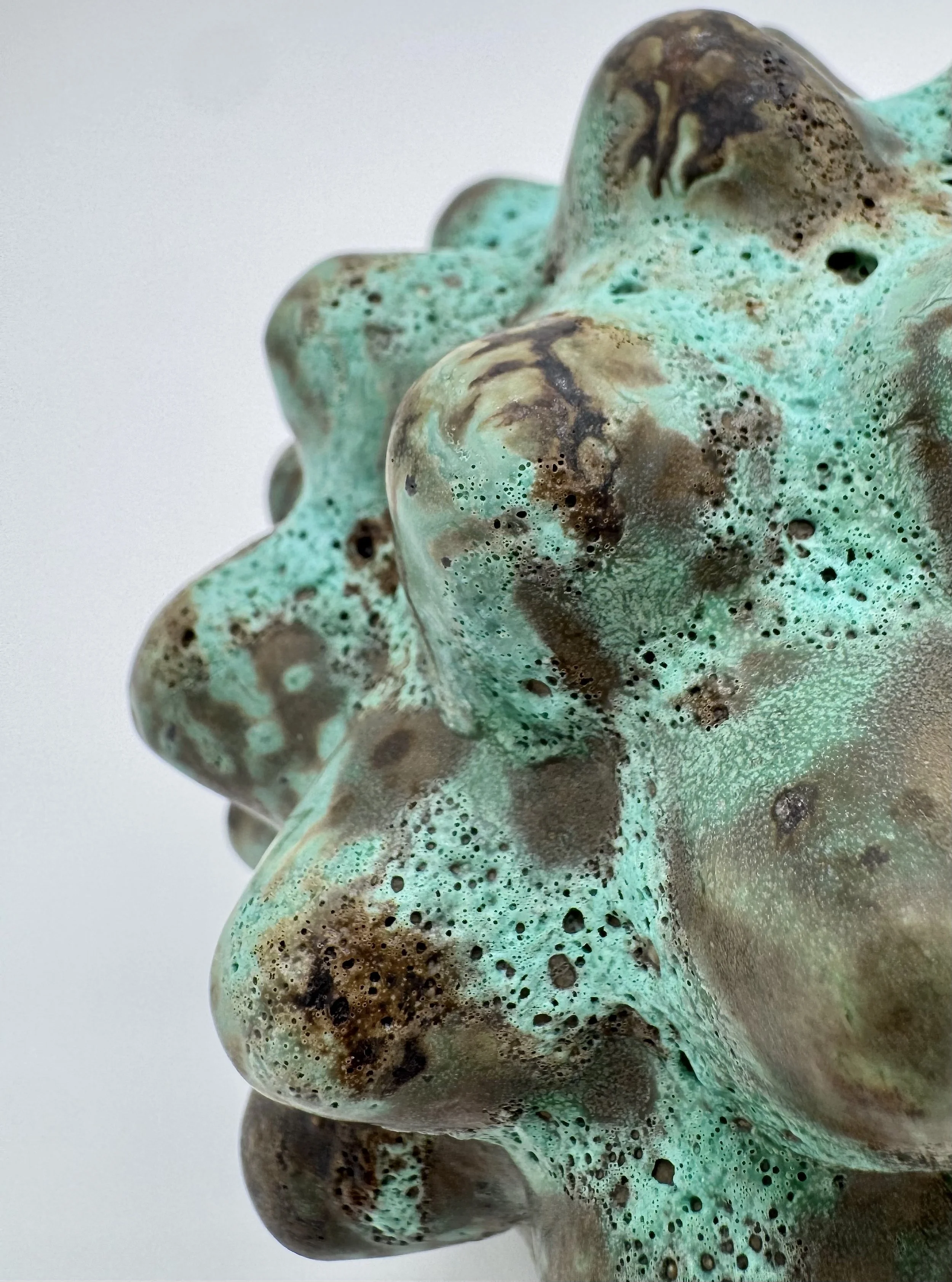 Close-up of a textured sculpture resembling a cluster of abstract, organic shapes with a mixture of turquoise, brown, and black colors, and small holes and bubbles on its surface.