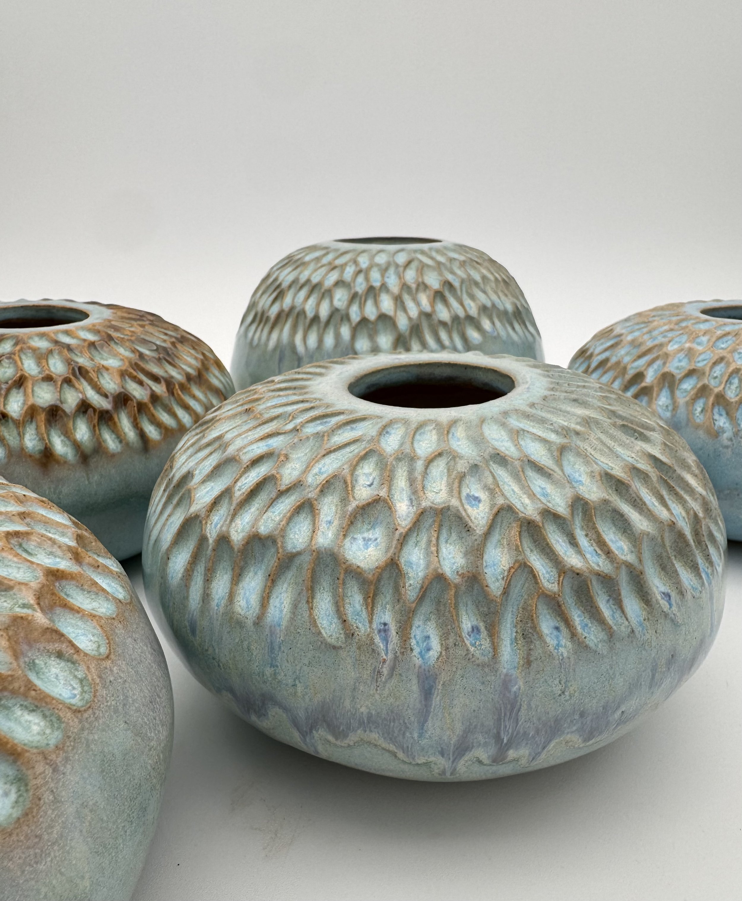 Close-up of multiple ceramic vases with textured, patterned surfaces in shades of cream, light blue, and brown, arranged on a white surface.