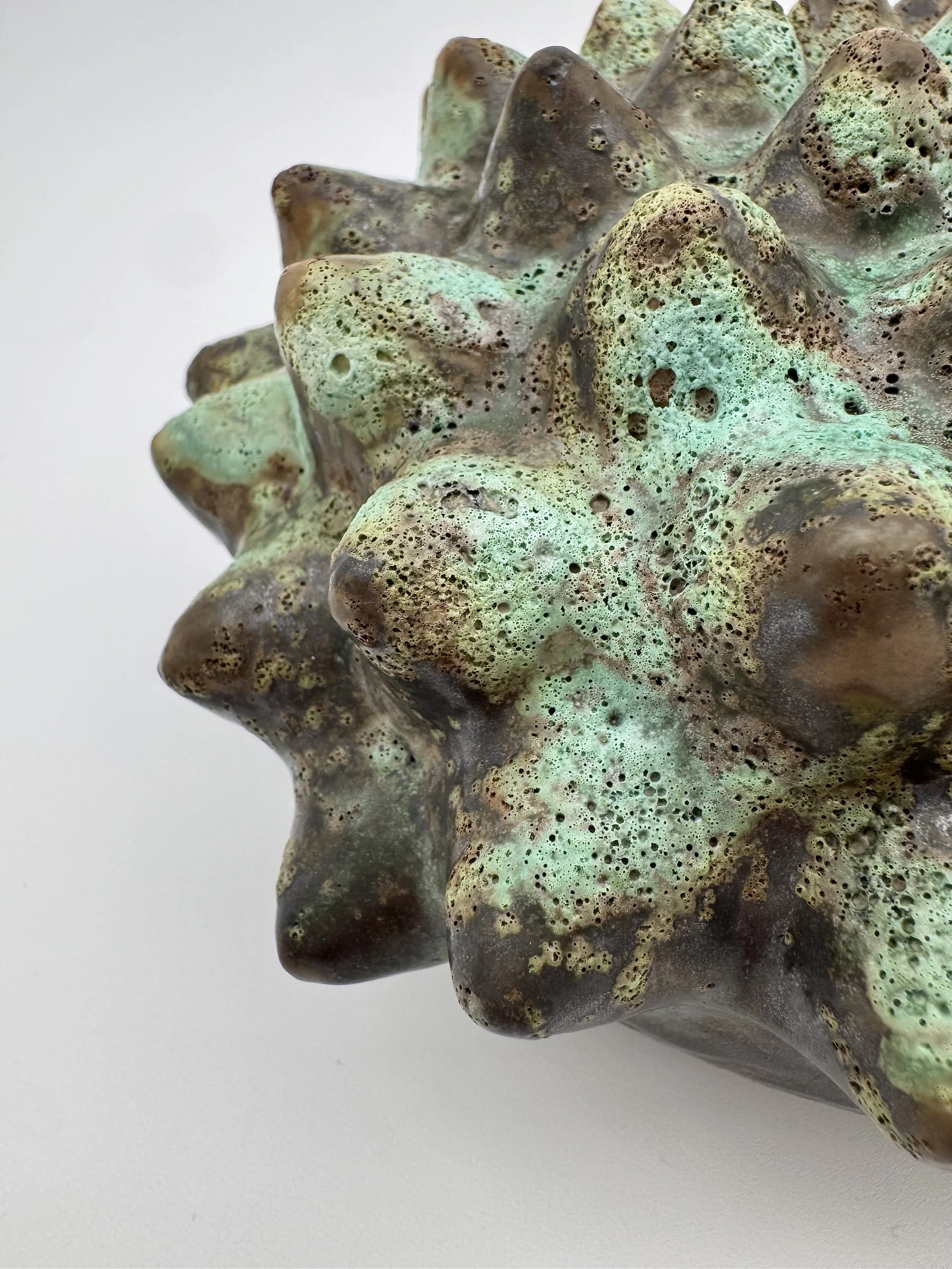 Close-up of a large, textured mineral or rock formation with green, brown, and black coloration and numerous small holes, set against a plain white background.