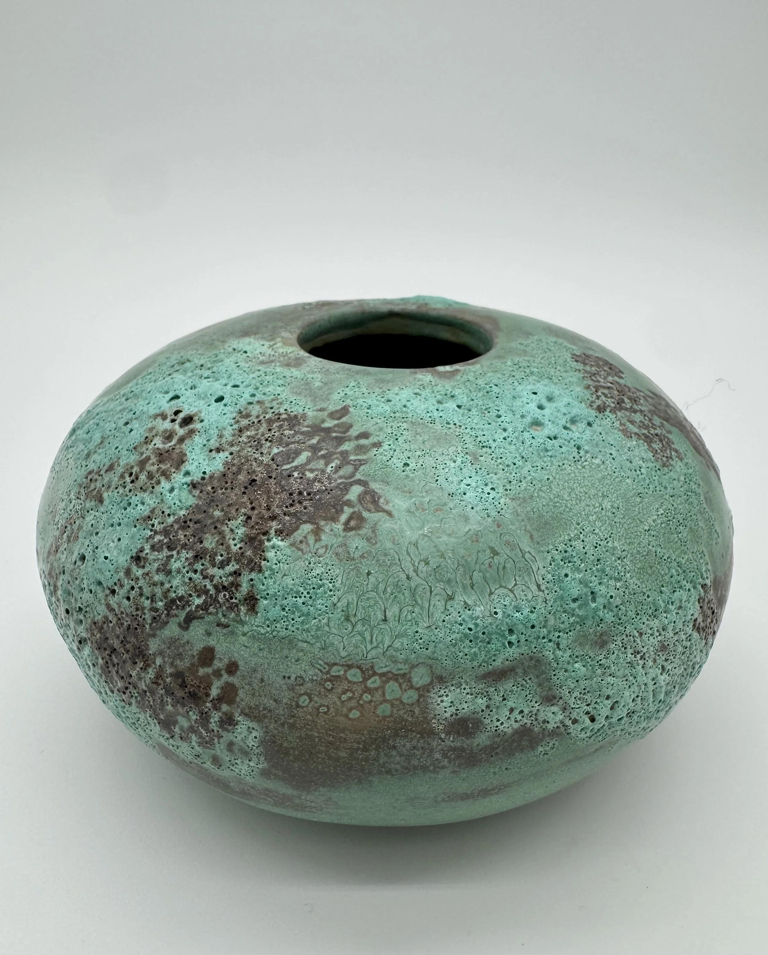 Close-up of a round, speckled turquoise ceramic vase with a narrow opening at the top, placed on a white surface against a plain white background.