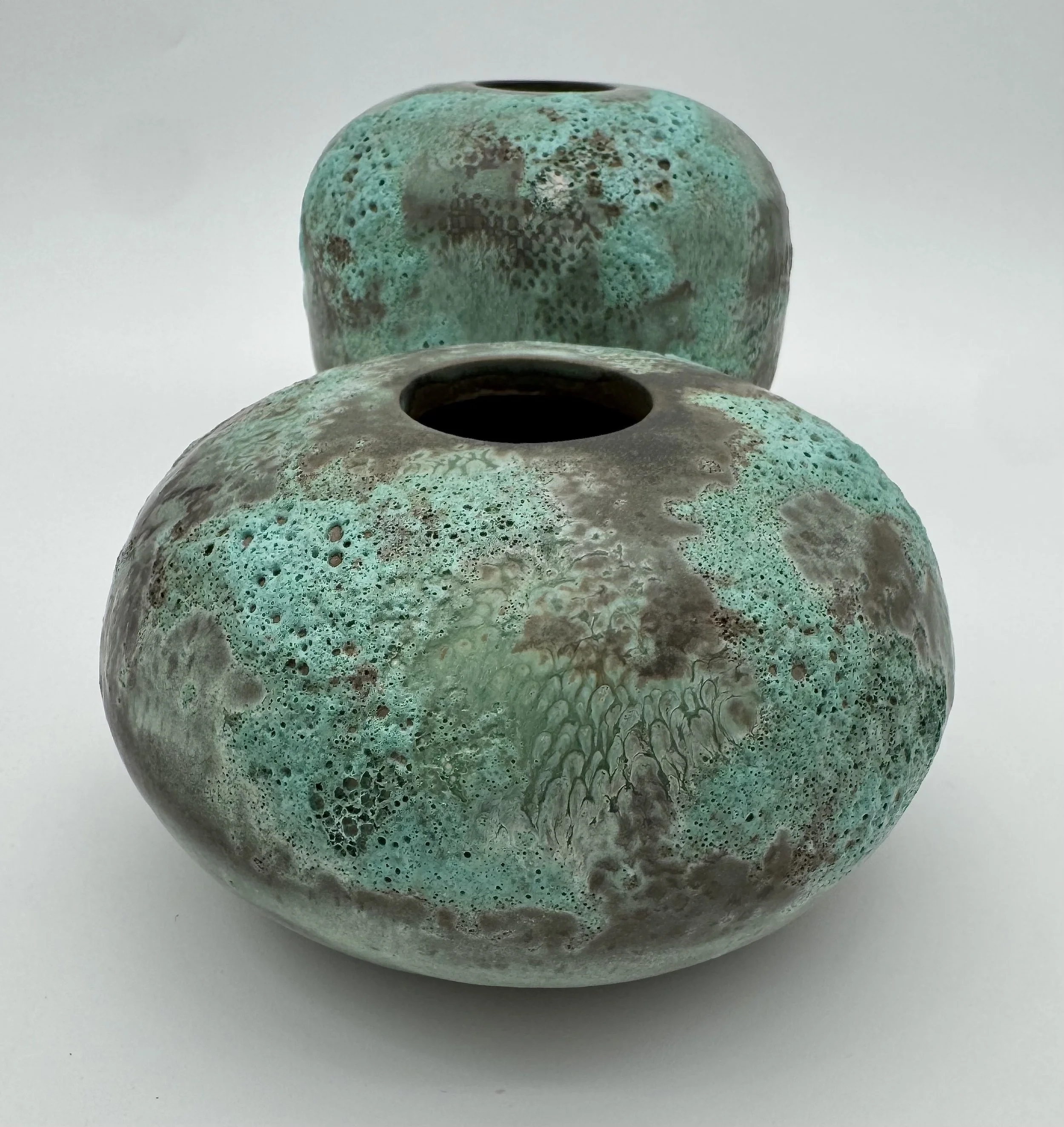 Two ceramic vases with textured, speckled green and brown surfaces, one partially visible in front of the other, against a plain white background.