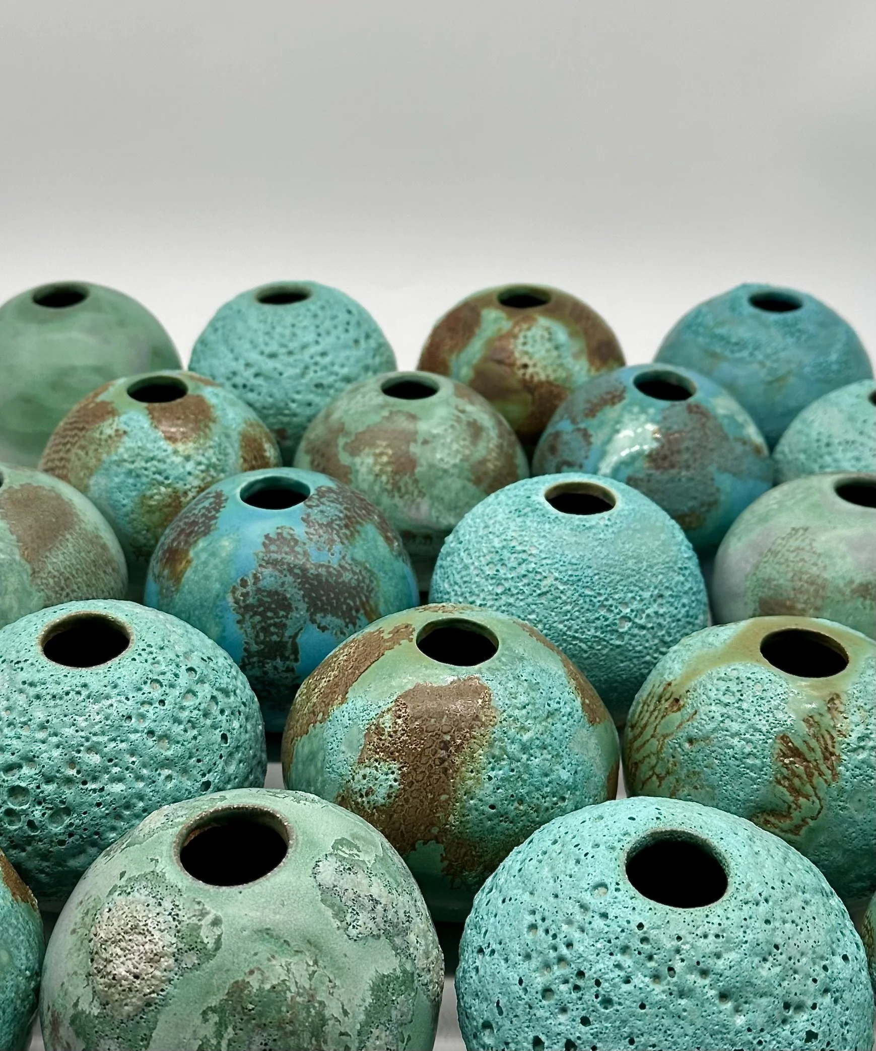 Close-up view of a collection of ceramic vases with textured surfaces in shades of teal, turquoise, and brown on a white background.
