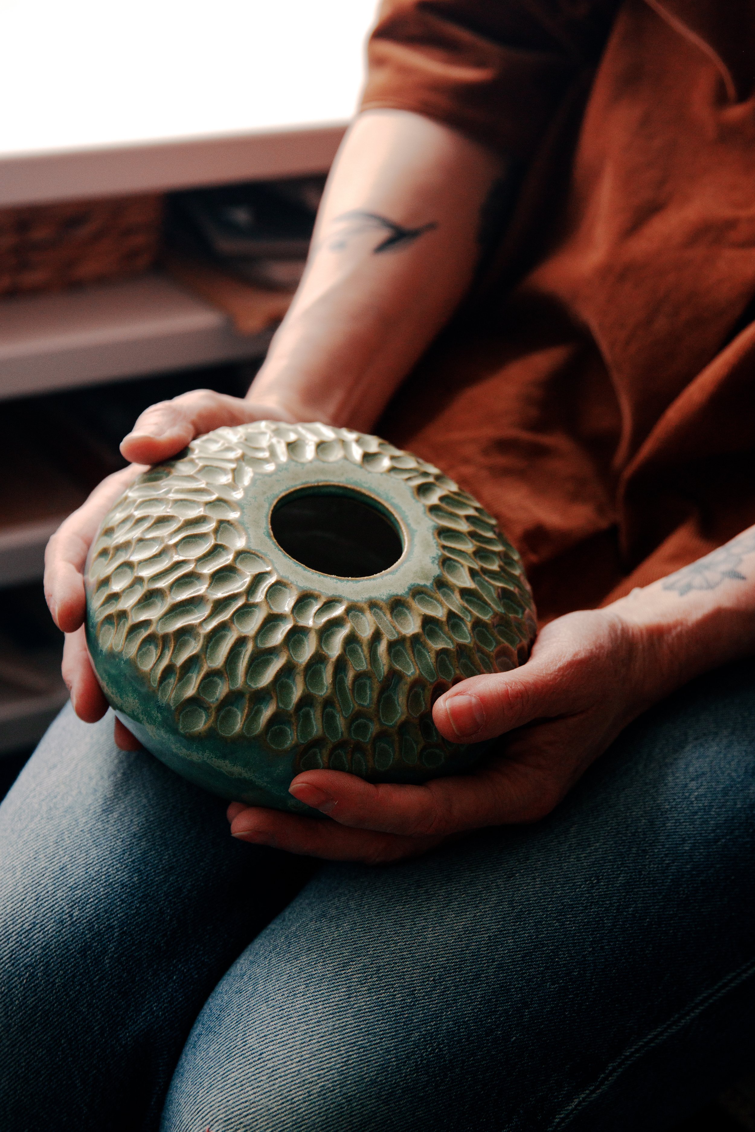 Person sitting with legs crossed, holding a ceramic decorative object with a textured pattern and a hollow center.