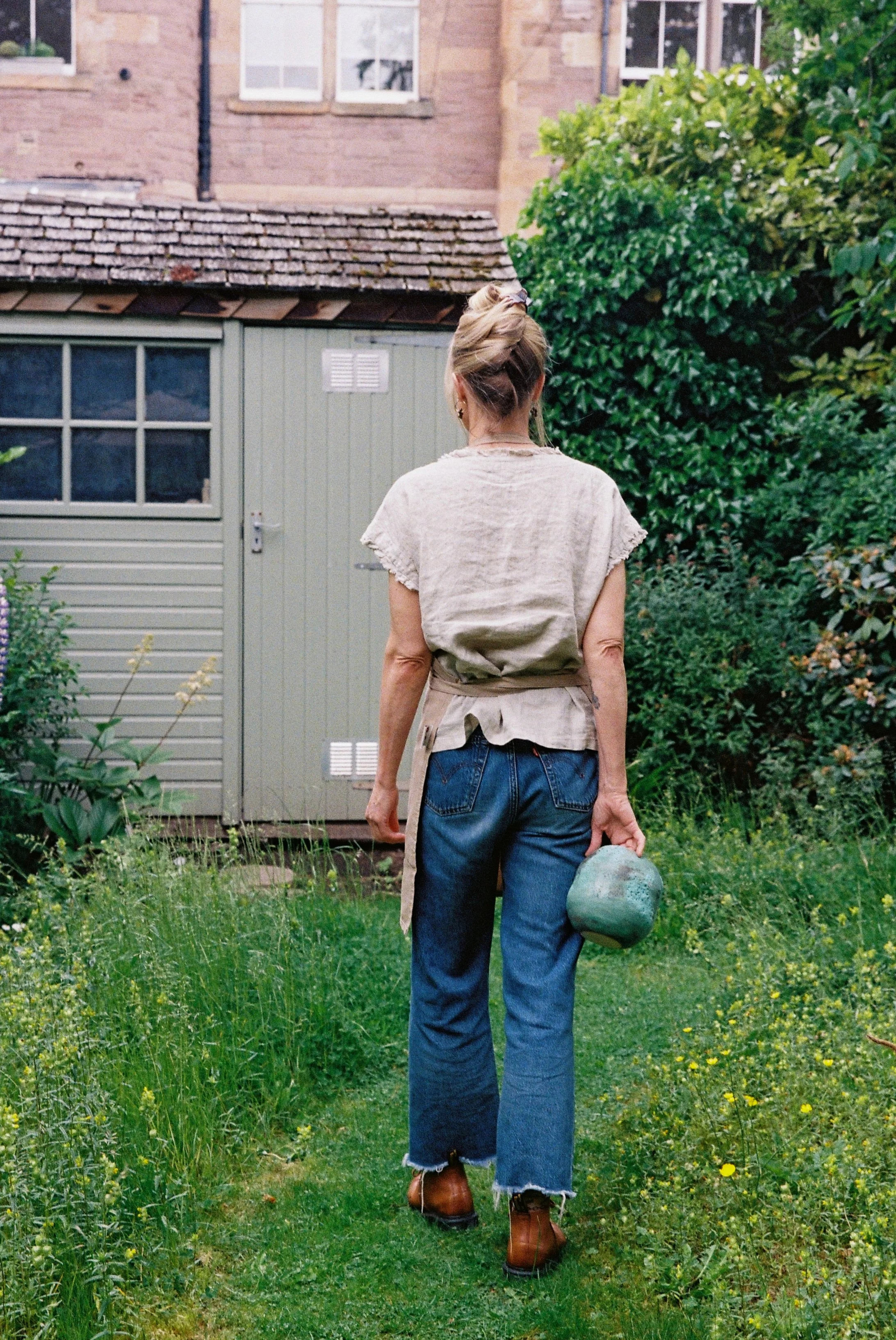 A woman walking away in a garden with green grass and bushes, holding a rock in her right hand, near a small shed and a brick house.