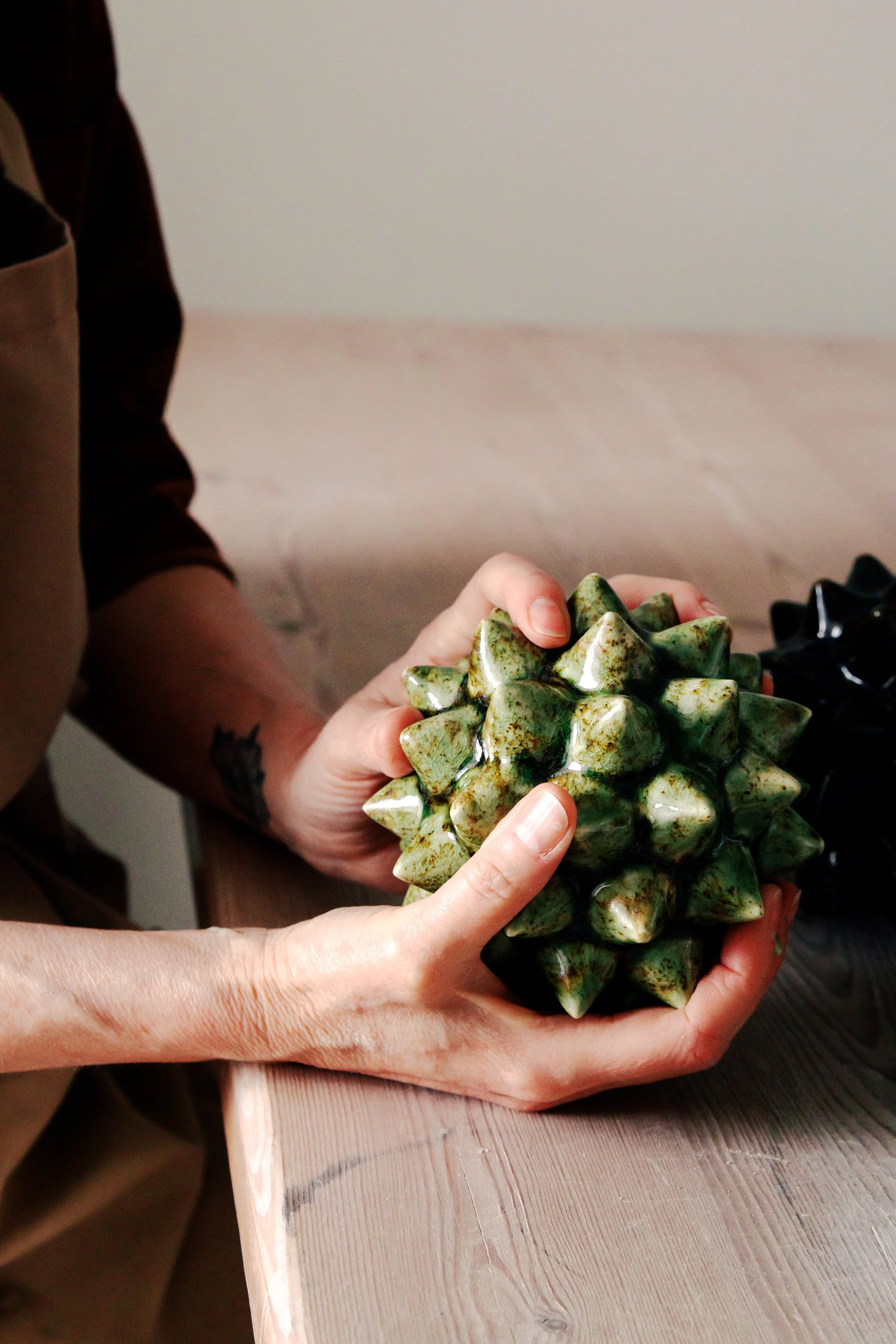 Person holding a green and white speckled, spiky ceramic art piece over a light wooden table.