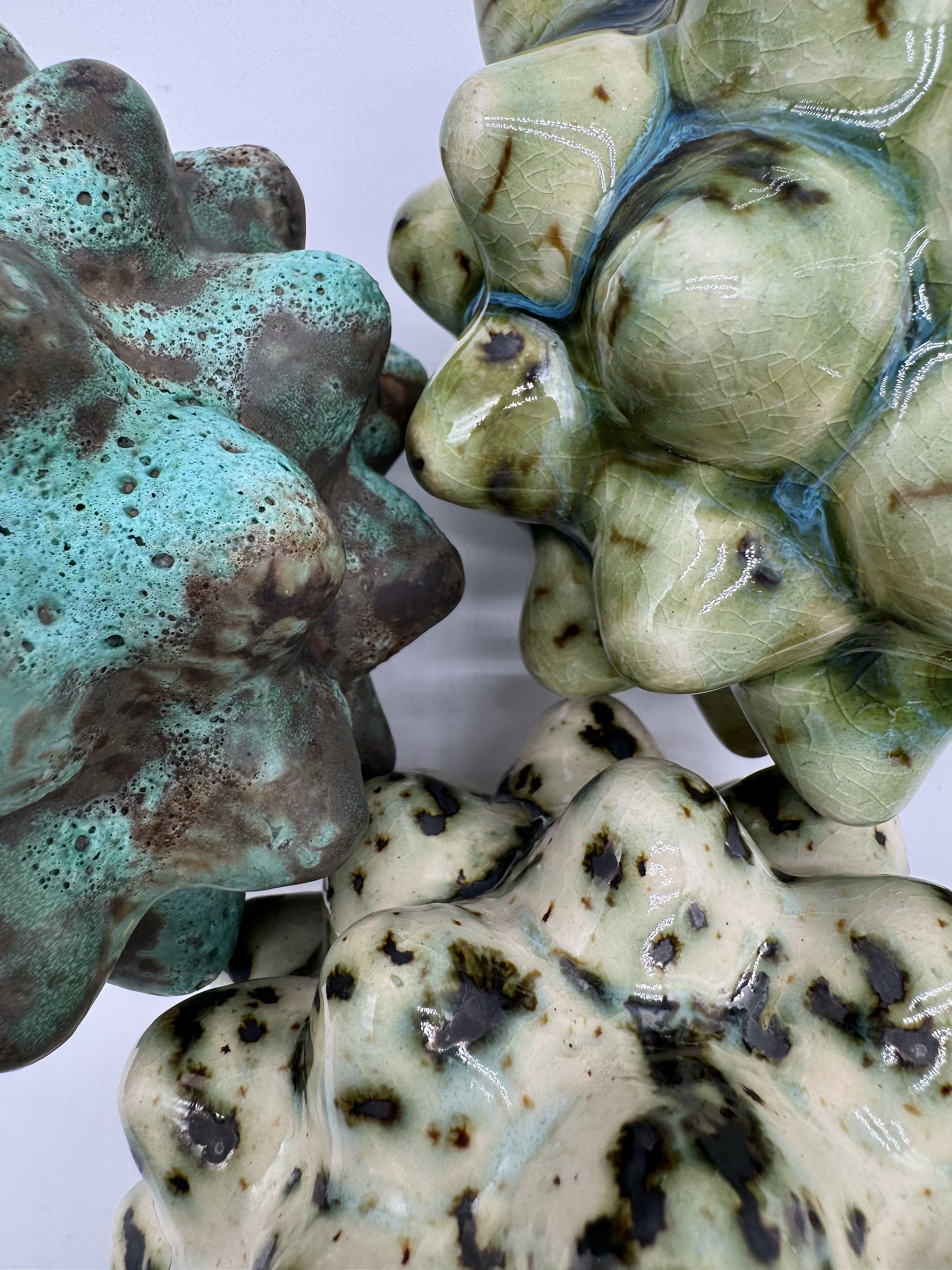 Close-up view of ceramic sculptures resembling abstract organic shapes in shades of green, brown, and off-white, with textured surfaces and crackle effects.