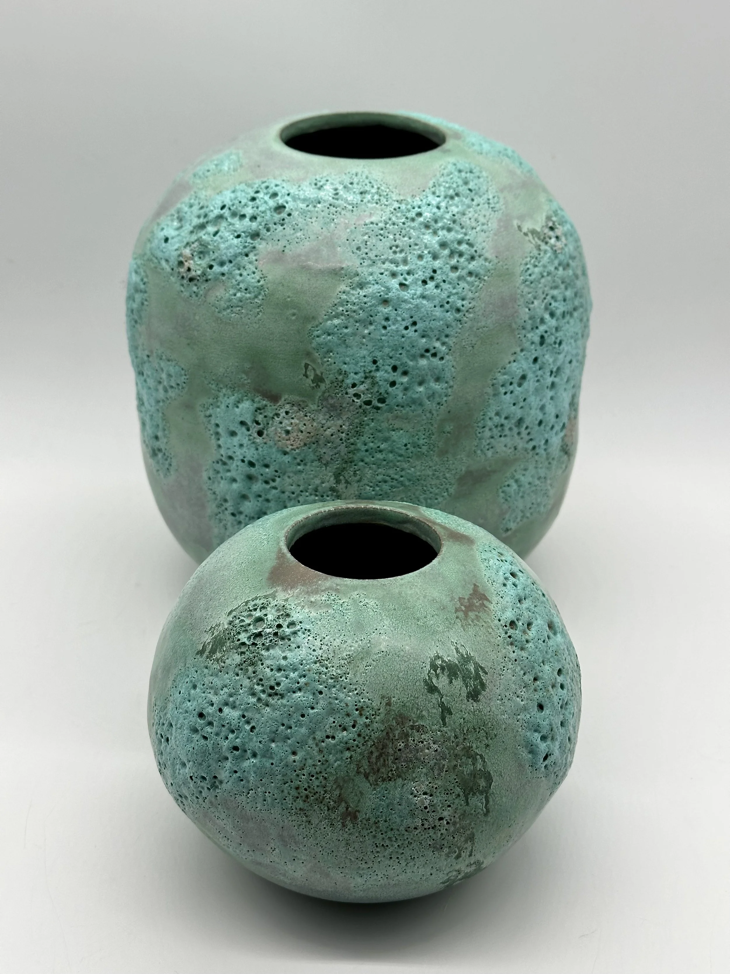 Two round ceramic vases with a bluish-green glaze, featuring textured, pockmarked surfaces and narrow openings at the top, placed on a white background.