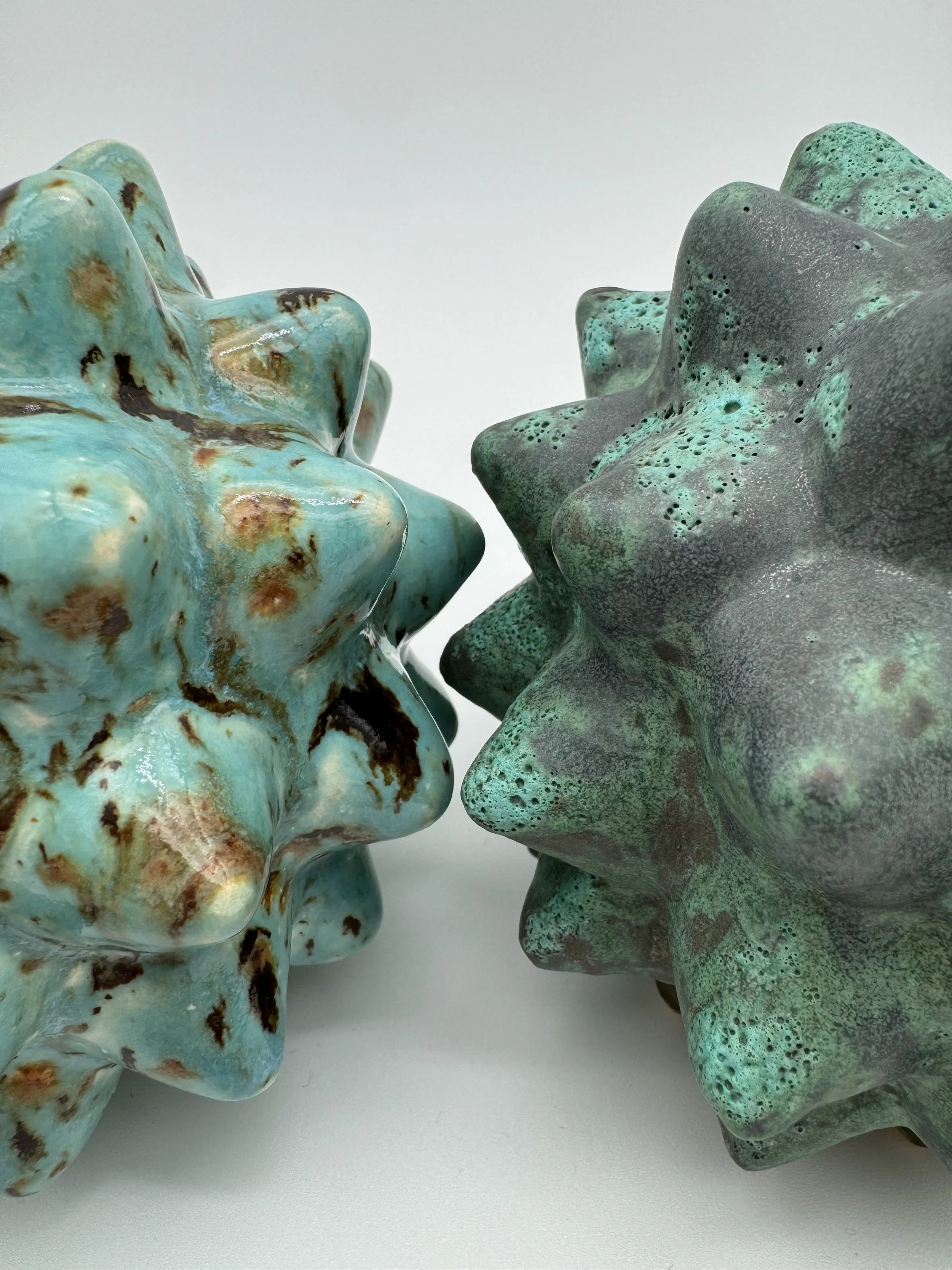 Close-up of two ceramic sculptures resembling abstract human faces with starfish-like textures, one in turquoise with brown spots, the other in dark green with blue accents, against a plain white background.