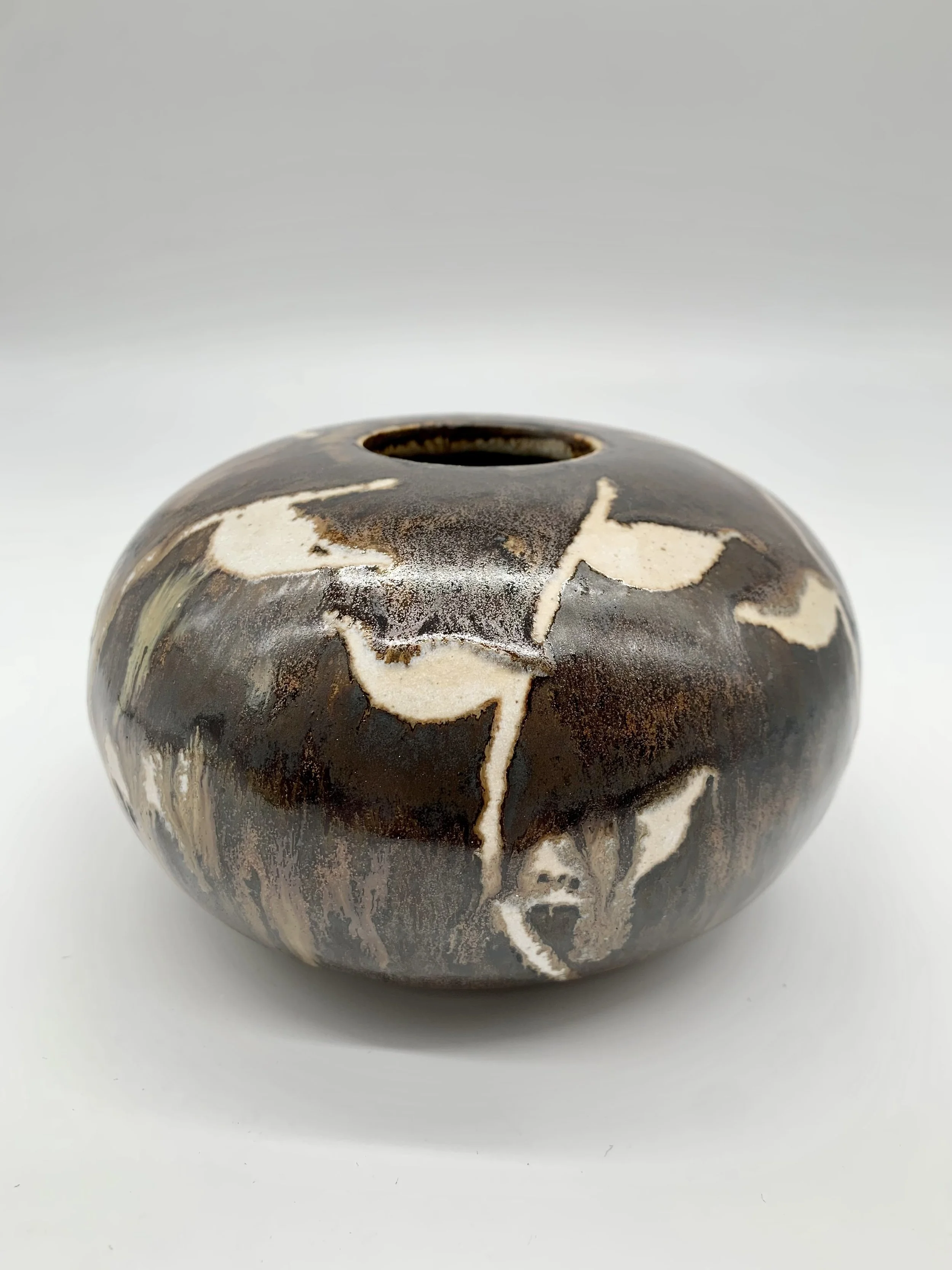 A round ceramic vase with a dark and cream marbled glaze, featuring a circular opening at the top, placed on a white surface.