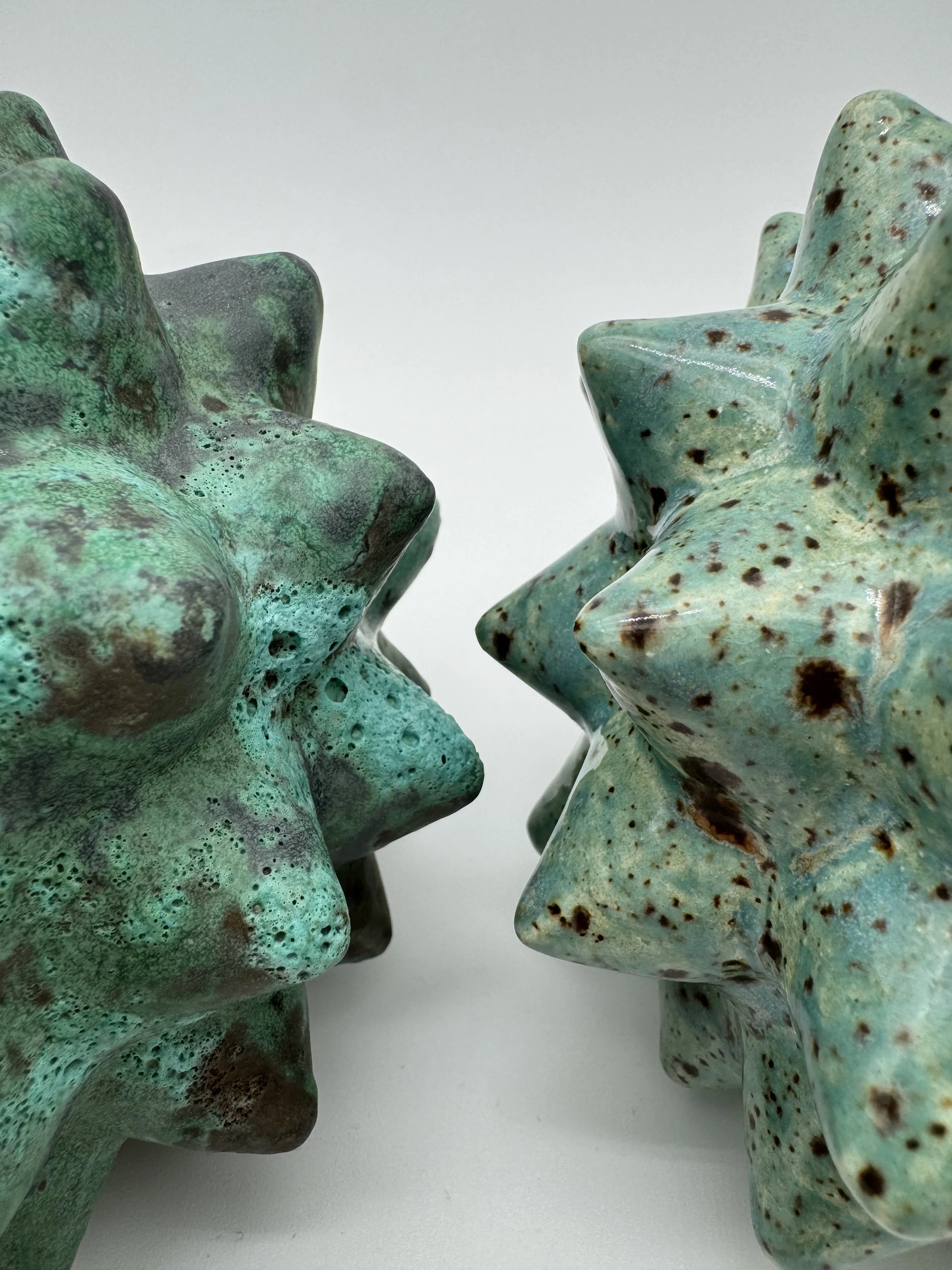 Two greenish ceramic starfish sculptures with a textured surface and brown speckles, placed on a white background.