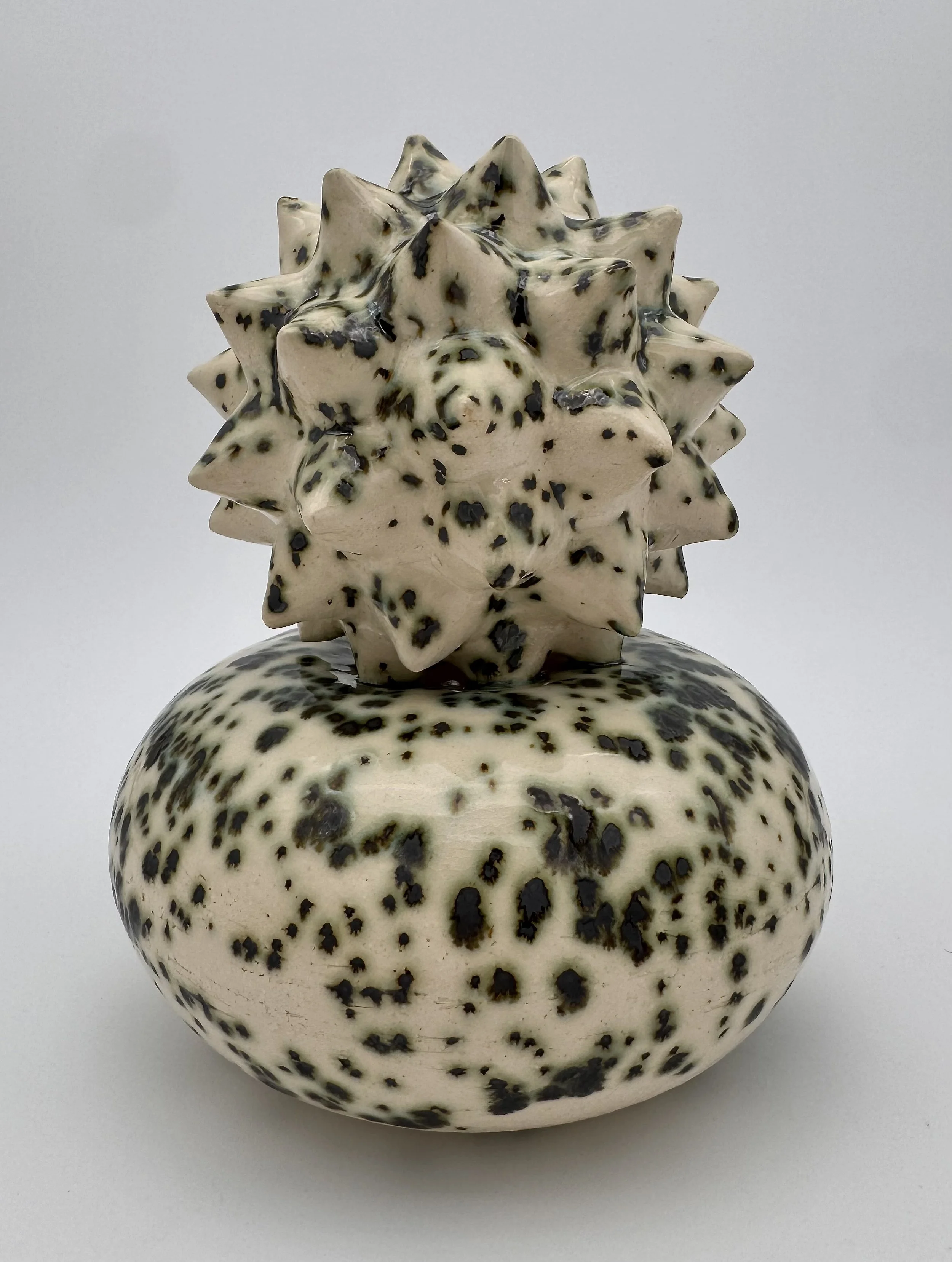 A decorative ceramic vase with a rounded base, featuring an intricate star-shaped top with multiple pointed spikes, decorated with black spots on a cream background.