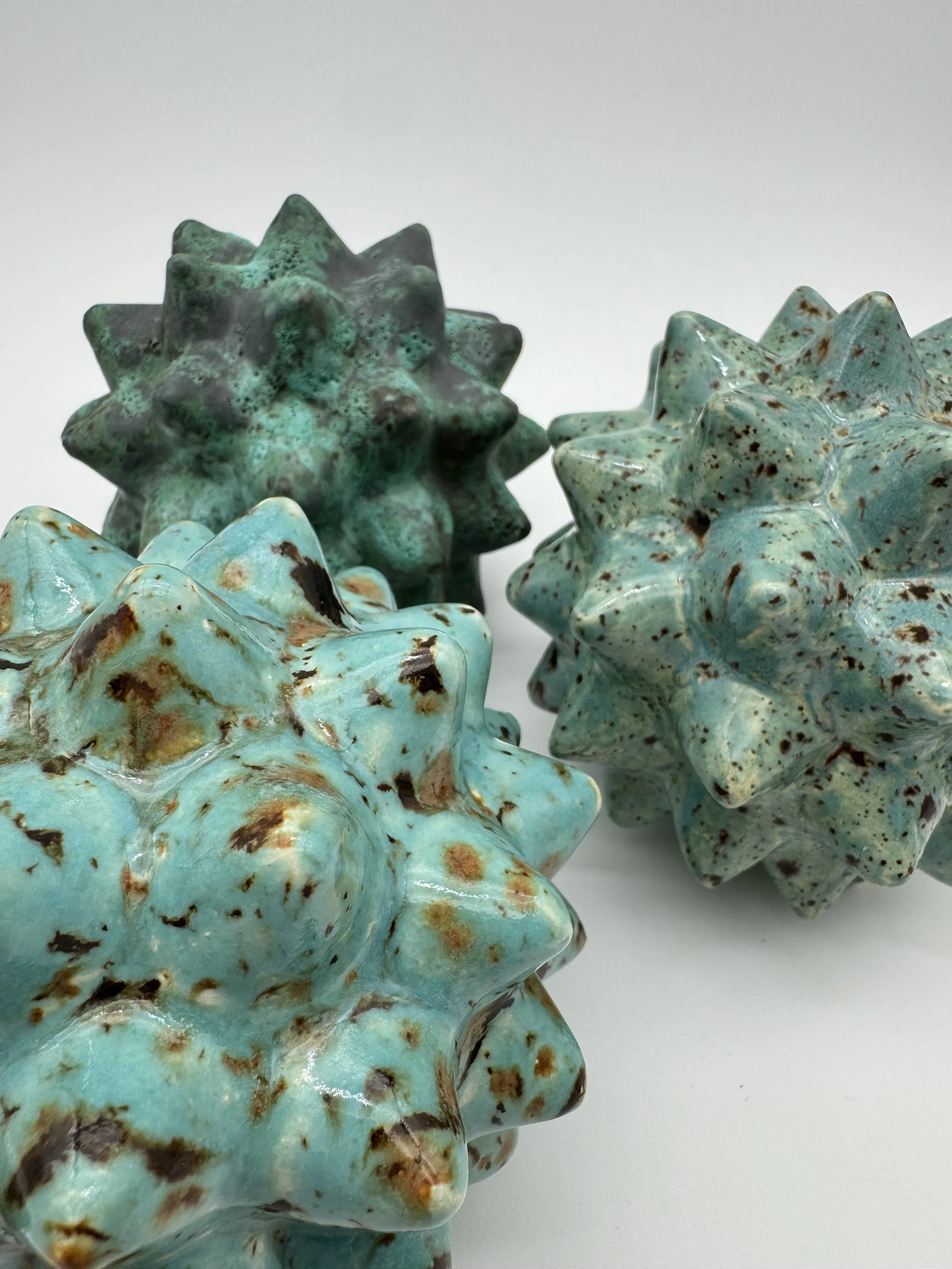 Close-up of three handcrafted ceramic star-shaped sculptures with a turquoise glaze and brown speckles, placed on a white background.