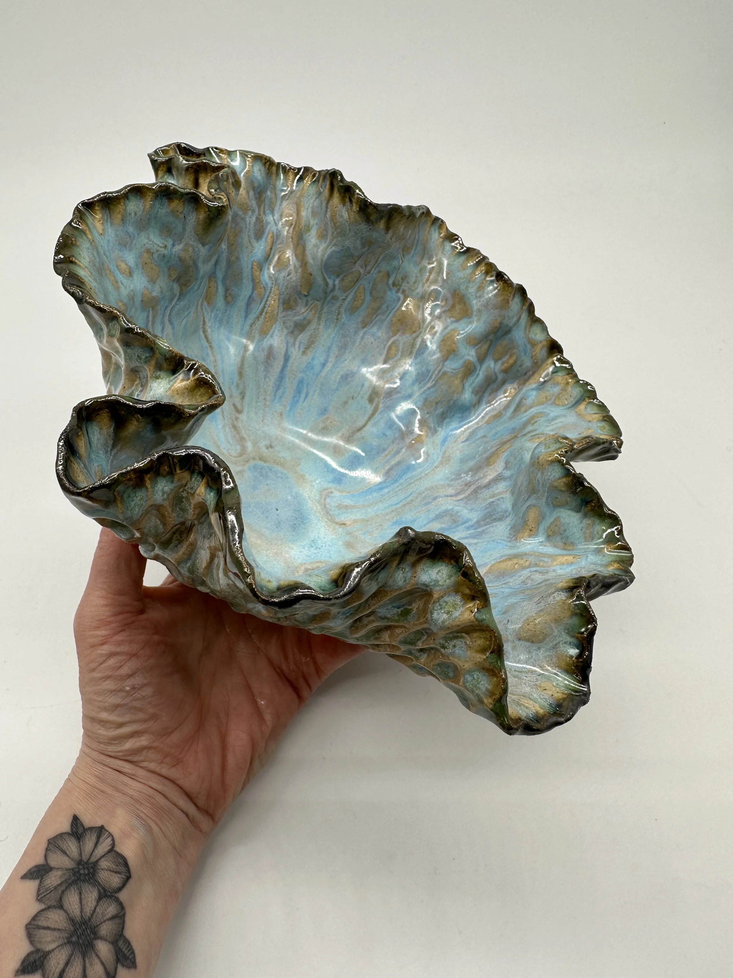 Close-up of a decorative ceramic leaf-shaped bowl with blue, brown, and green glazing, being held by a hand with tattoos on the wrist.