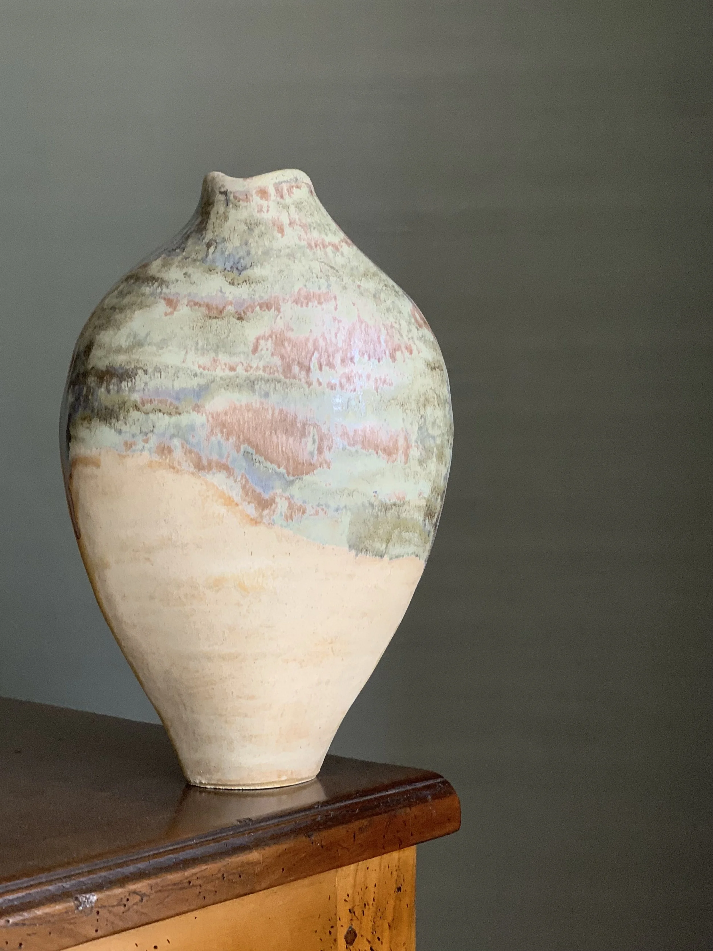 A ceramic vase with a rounded body and a narrow neck, featuring pastel-colored, abstract patterns, placed on a wooden surface against a plain background.