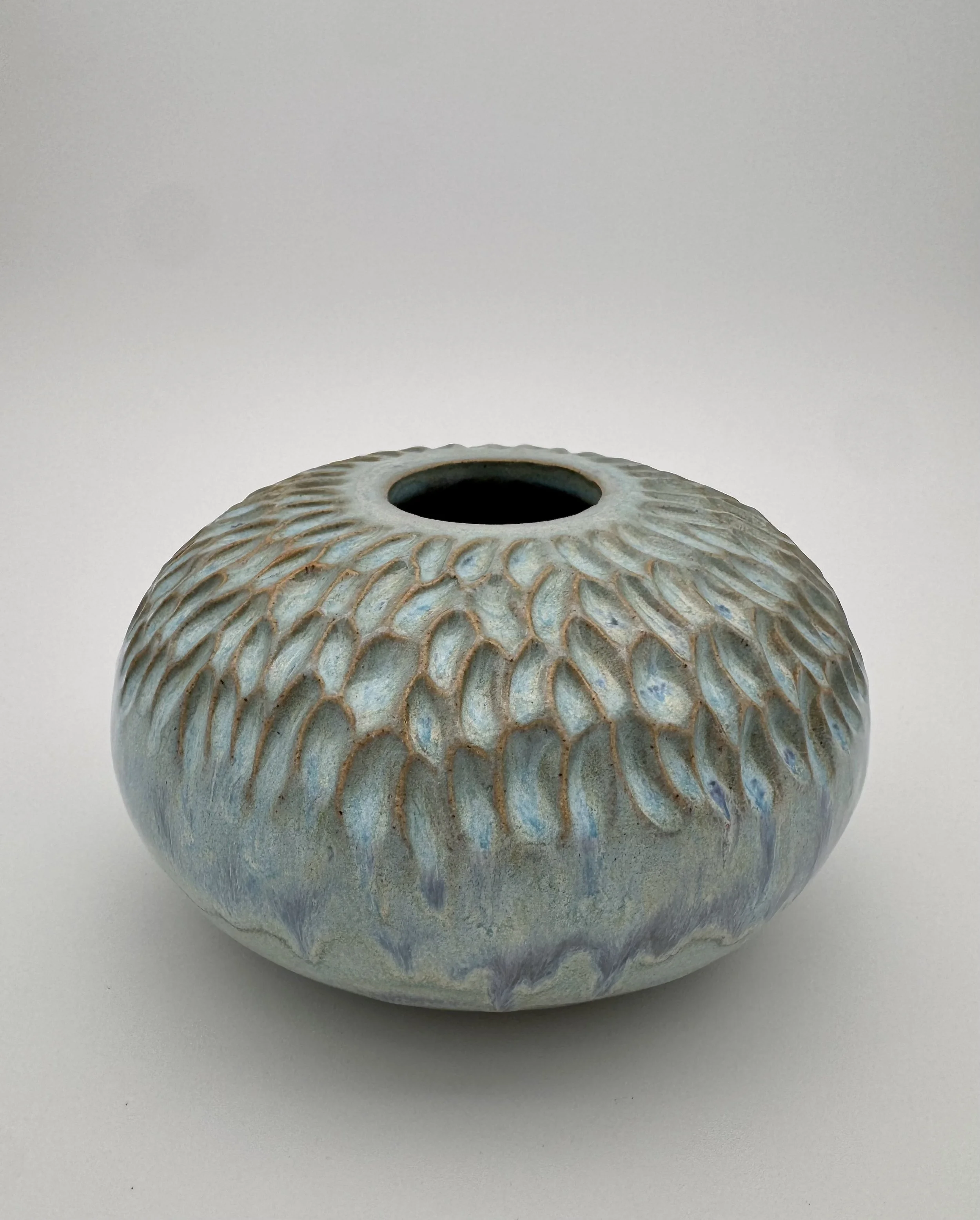 A ceramic vase with a rounded shape and a textured pattern of raised scales, painted in shades of pale blue and beige, sitting on a plain white surface.