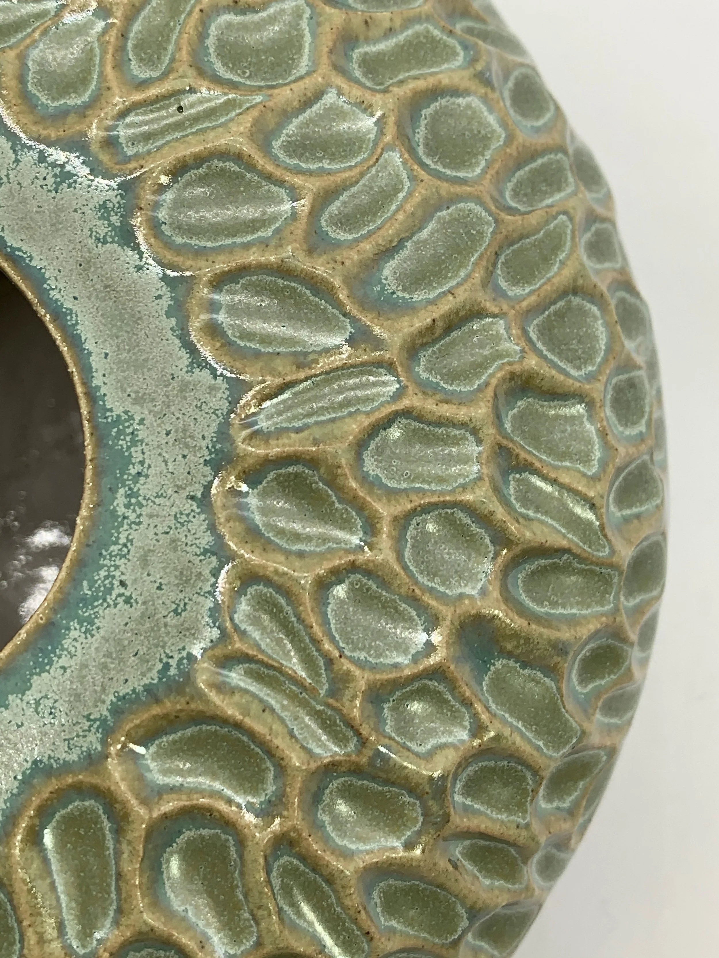 Close-up of a ceramic object with a textured pattern of raised ovals outlined in a beige color. The ovals are filled with a light greenish glaze, and the edges have a metallic, iridescent sheen. The background of the object is a speckled, bluish-gree