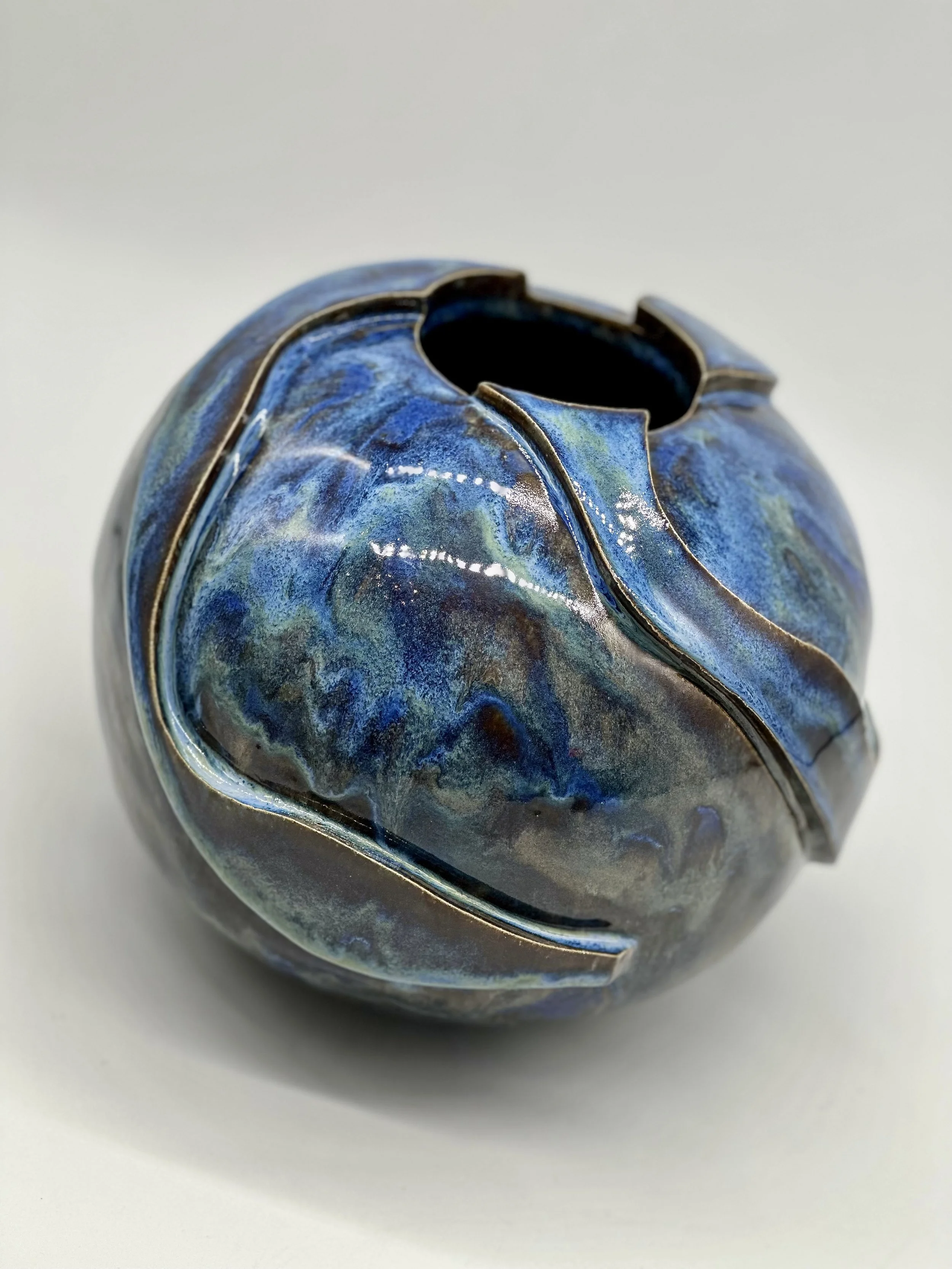 Close-up of a ceramic vase with a blue, green, and black abstract glaze pattern, viewed from above on a white surface.