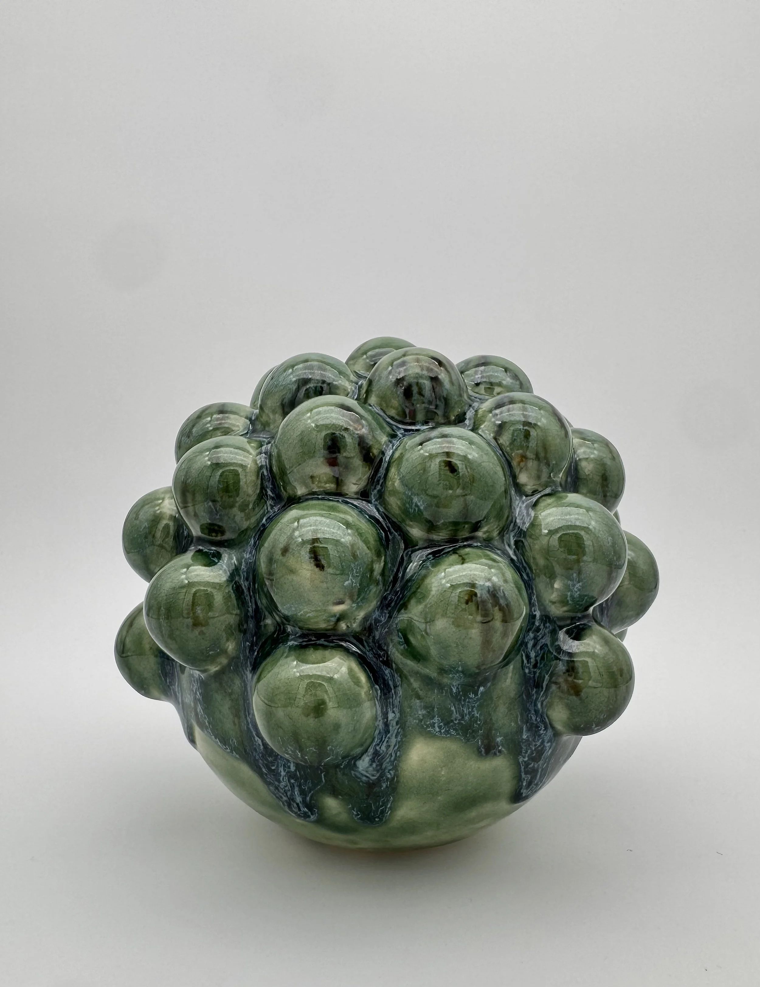 A glass art sculpture resembling a cluster of green grapes with a textured, multicolored surface placed on a plain white surface.