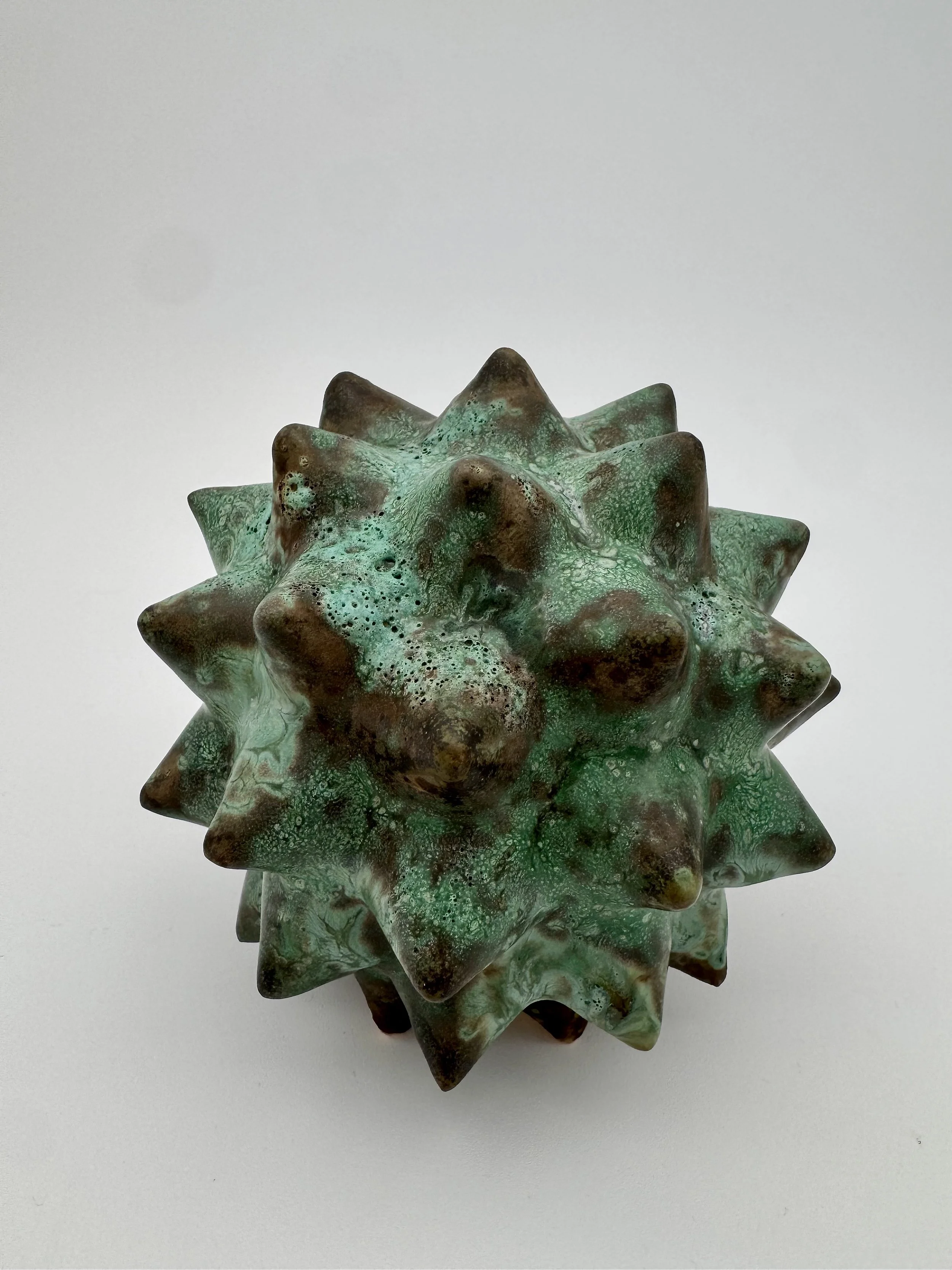 Close-up of a green and brown spiked ceramic sculpture against a plain background.