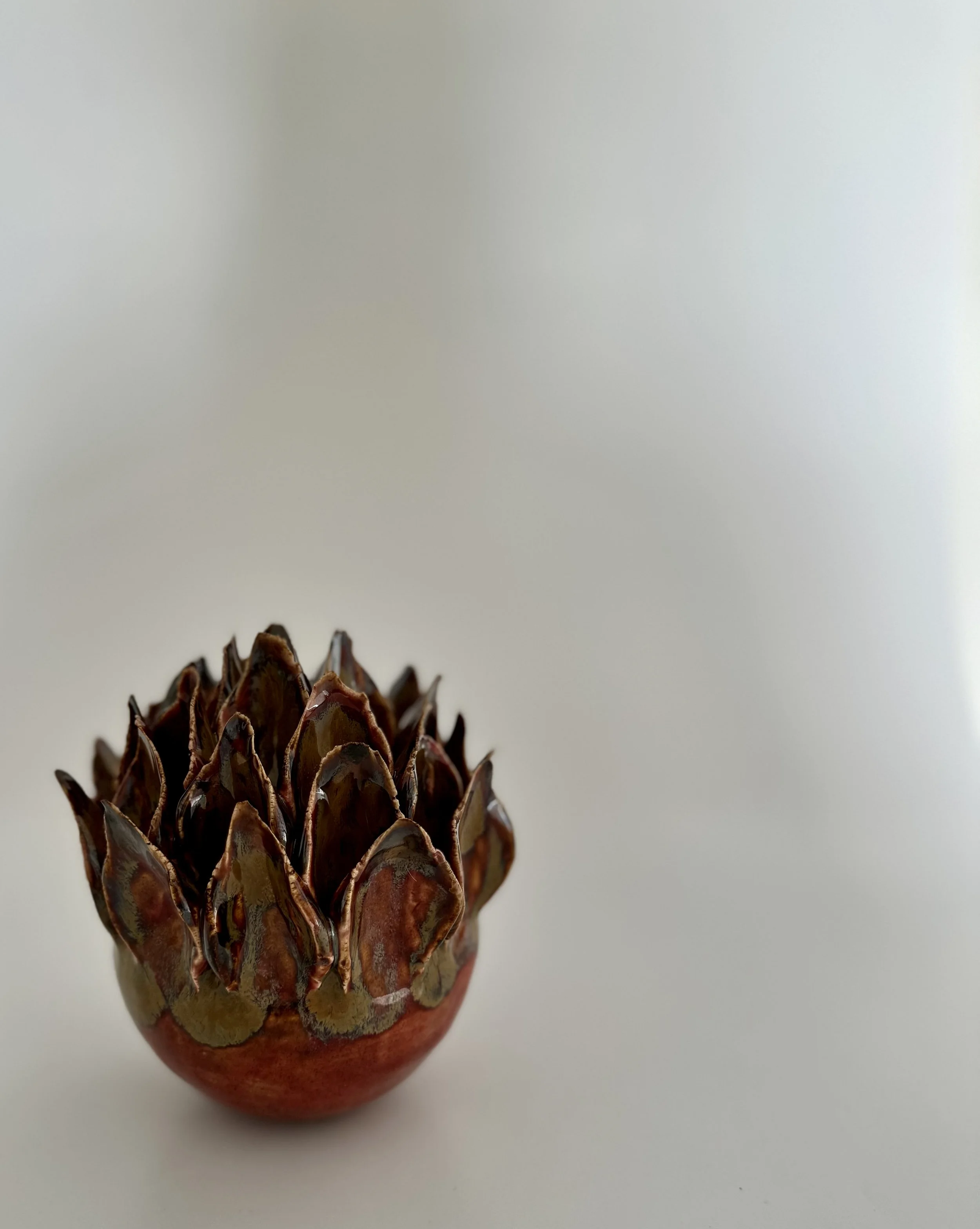 Close-up of a decorative object resembling a pinecone, made from metallic or ceramic material with a shiny, iridescent finish, placed on a plain white surface.