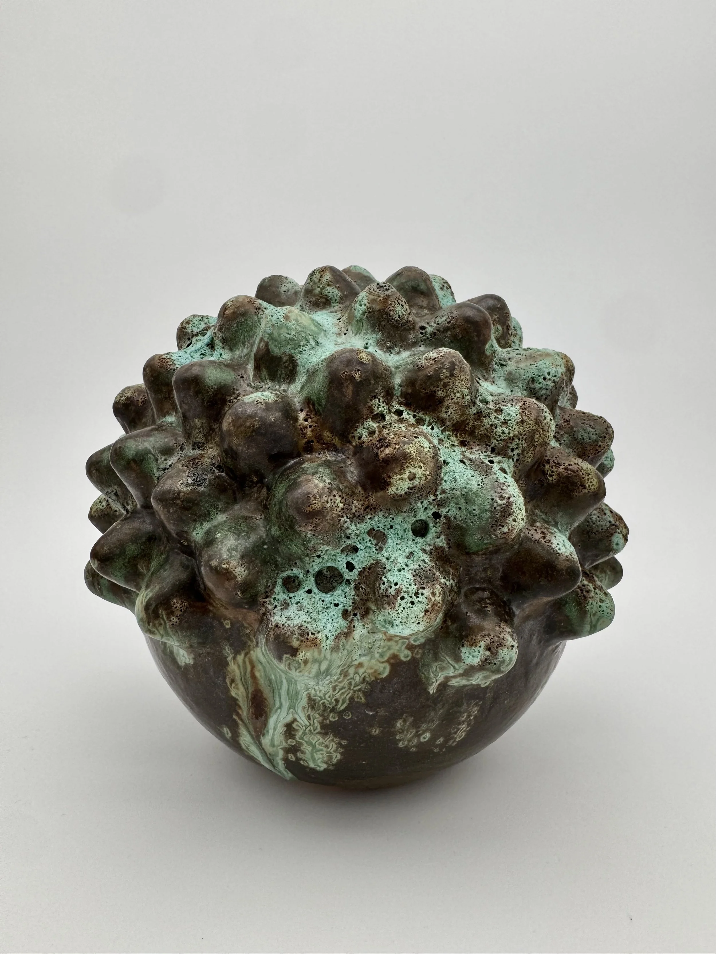 A ceramic art piece with a rounded shape, covered in textured bumps and a glaze with green, brown, and cream colors, placed on a white background.