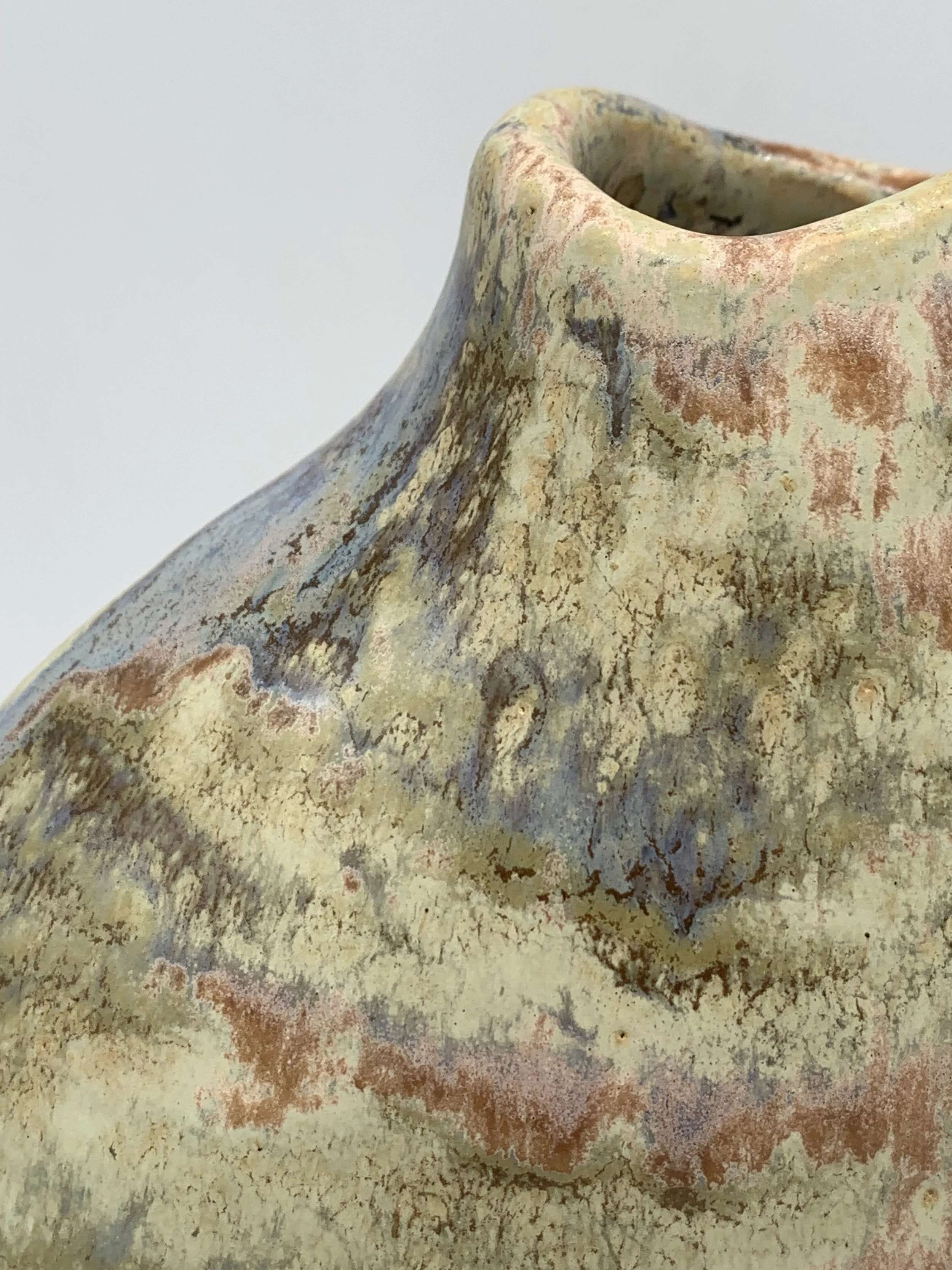 Close-up of a textured ceramic vase with a flared, round opening, showcasing a weathered, multicolored glaze with earthy tones of beige, brown, gray, and hints of red.