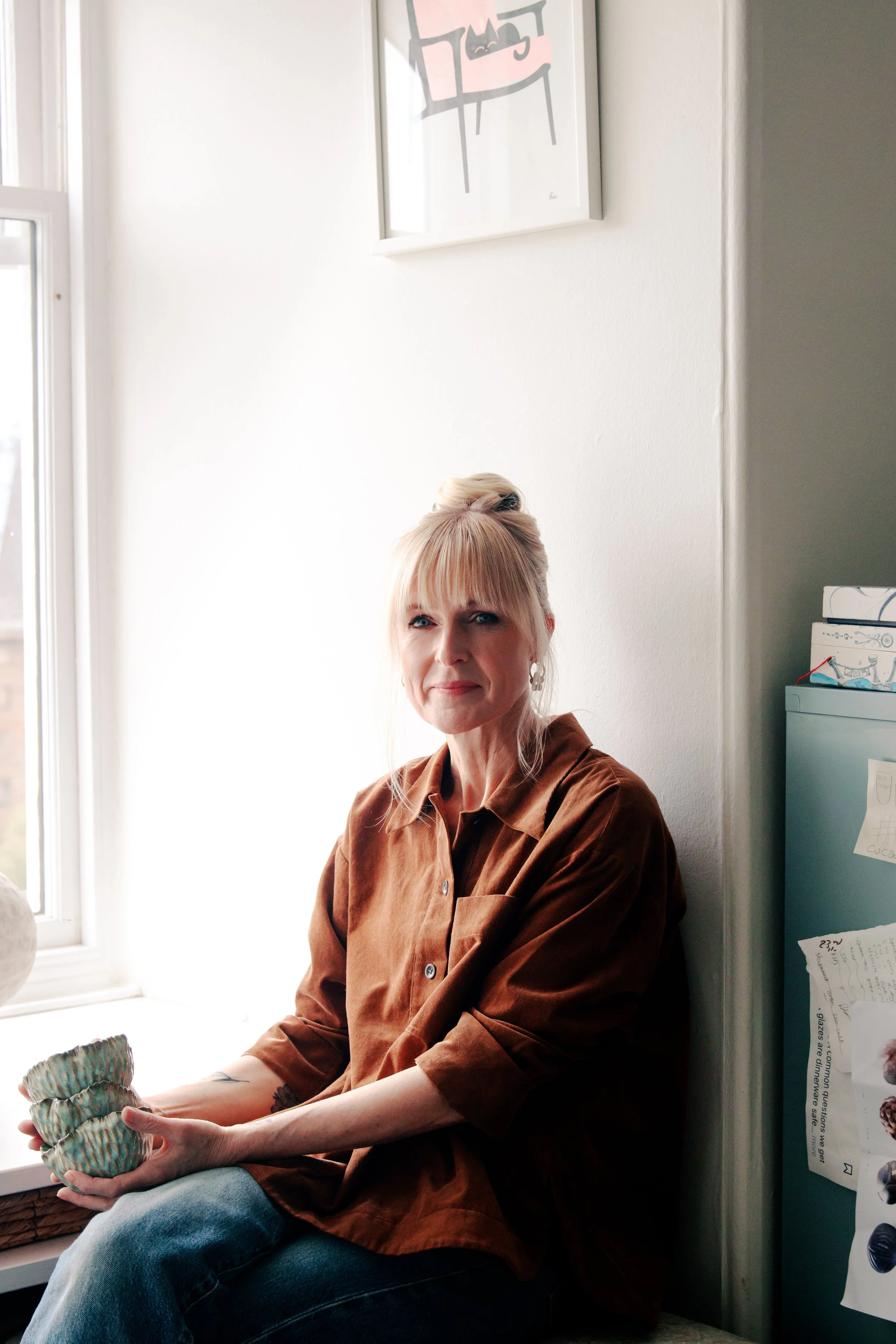 A woman with blonde hair tied in a bun, wearing a brown shirt, sitting by a window holding a woven basket.