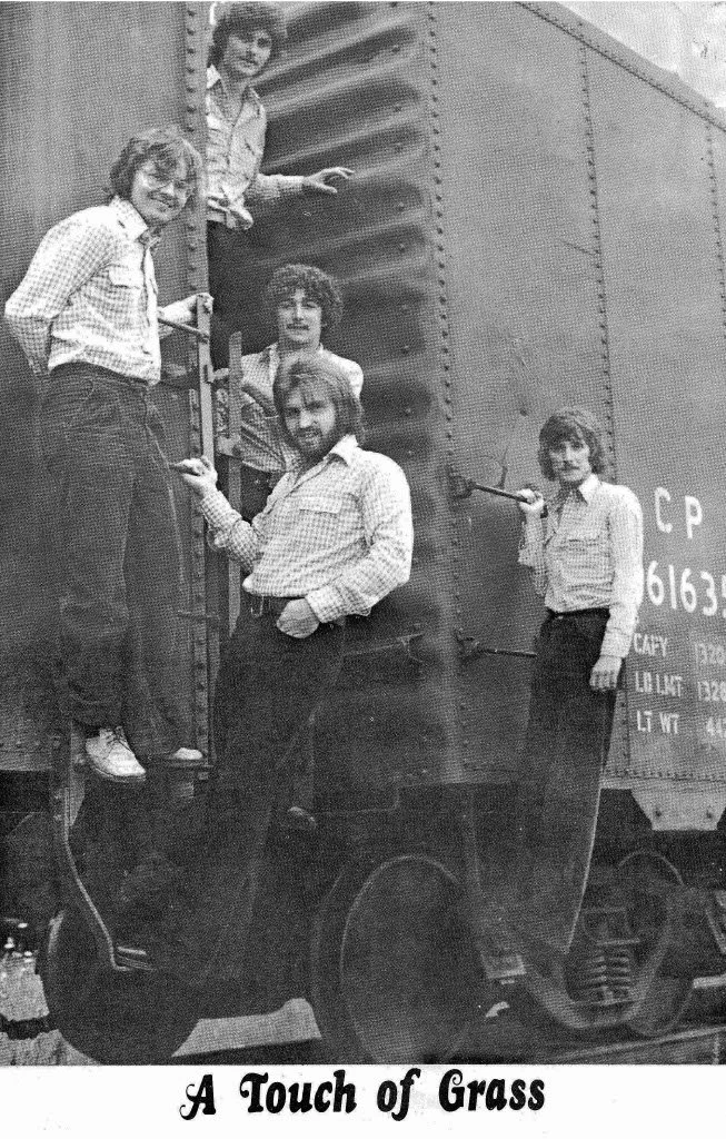 Black and white photograph of five young men climbing on a train car. They are wearing casual clothes, smiles, and posing. The text at the bottom reads "A Touch of Grass".
