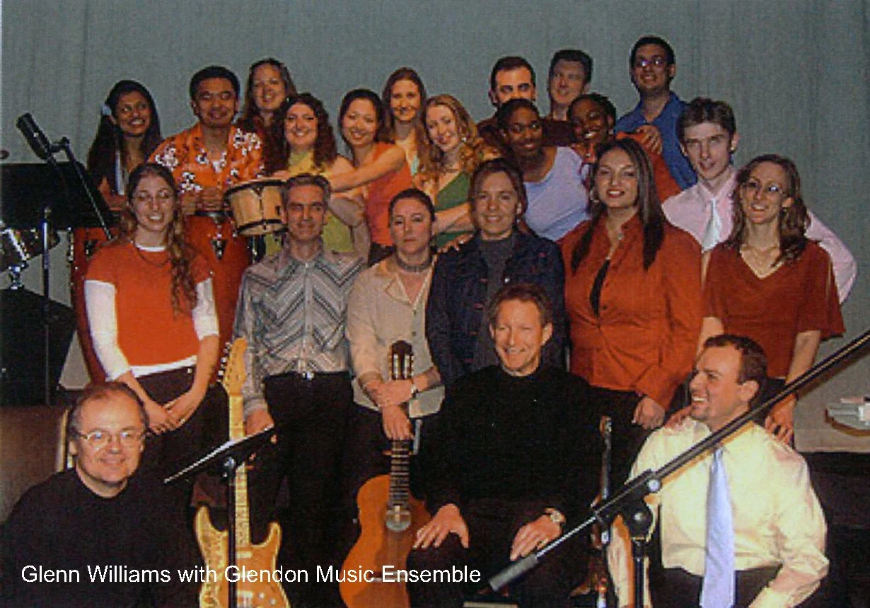 Group photo of Glenn Williams with Glendon Music Ensemble, with men and women of various ages, some holding musical instruments like guitars and tambourines, in a room with a plain background.