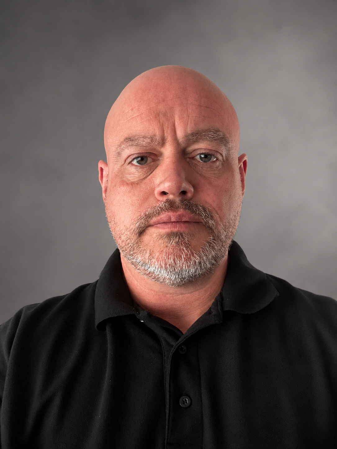 A bald man with a gray beard and mustache wearing a black collared shirt, looking serious against a gray background.