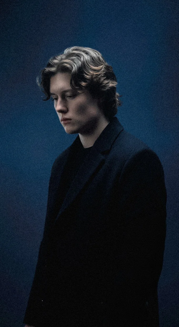 A young man with wavy, shoulder-length hair, wearing a dark blazer and shirt, standing against a textured blue background, looking downward with a contemplative expression.