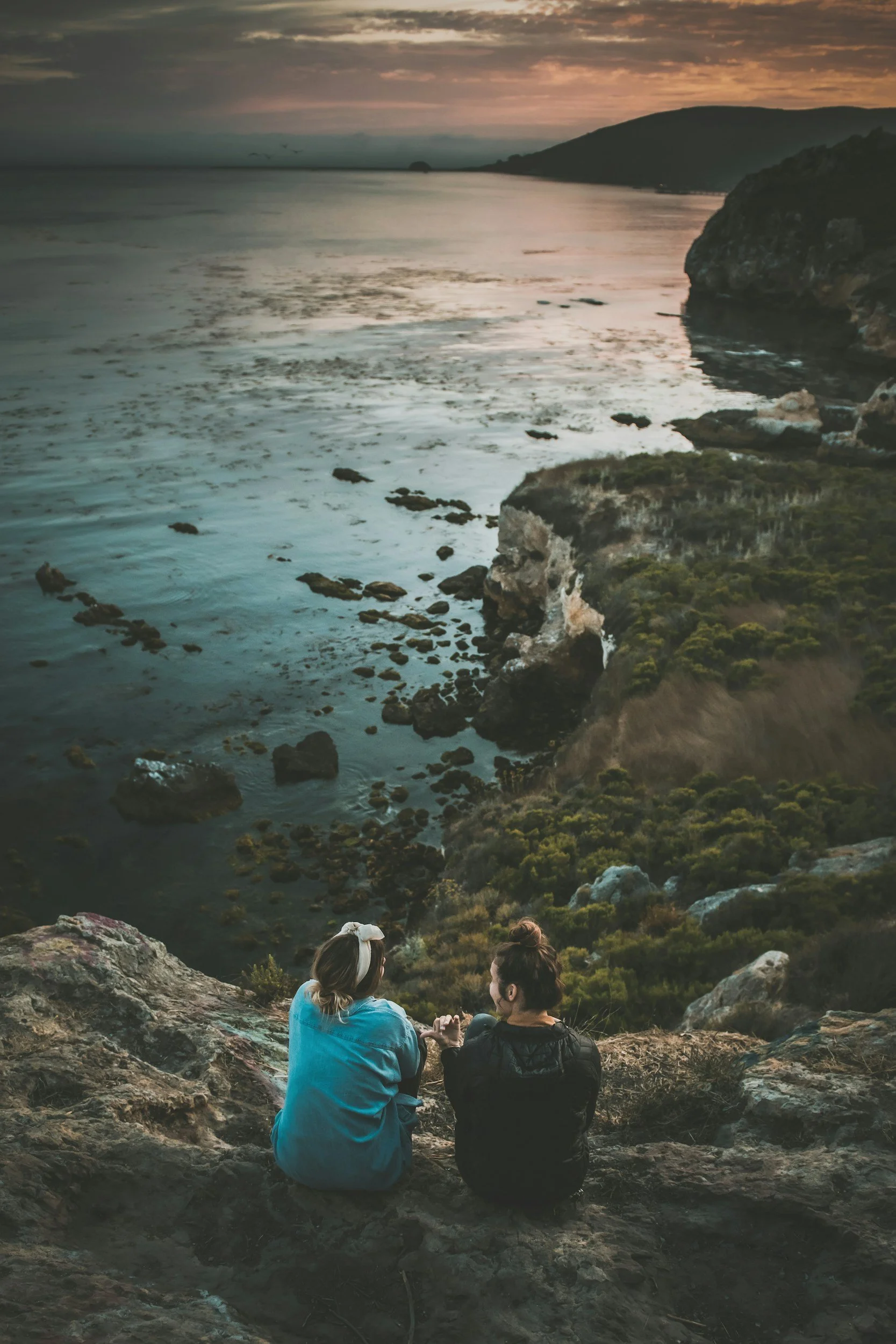 Two women sitting on rocks by the coast during sunset, with cliffs and water in the background.