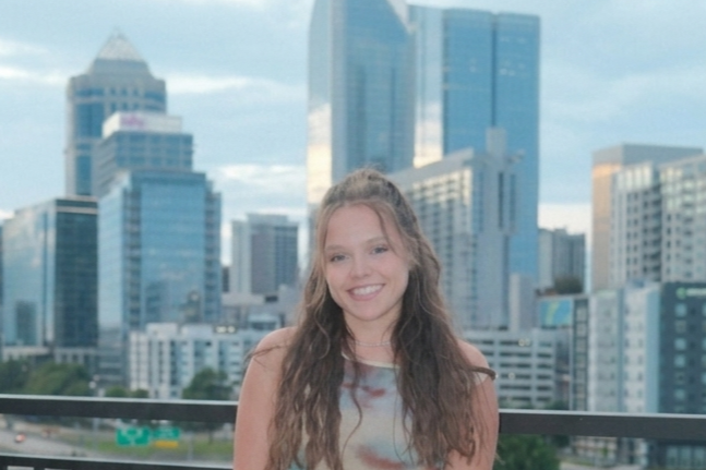 Smiling young woman with long, curly hair standing in front of a cityscape with tall buildings.