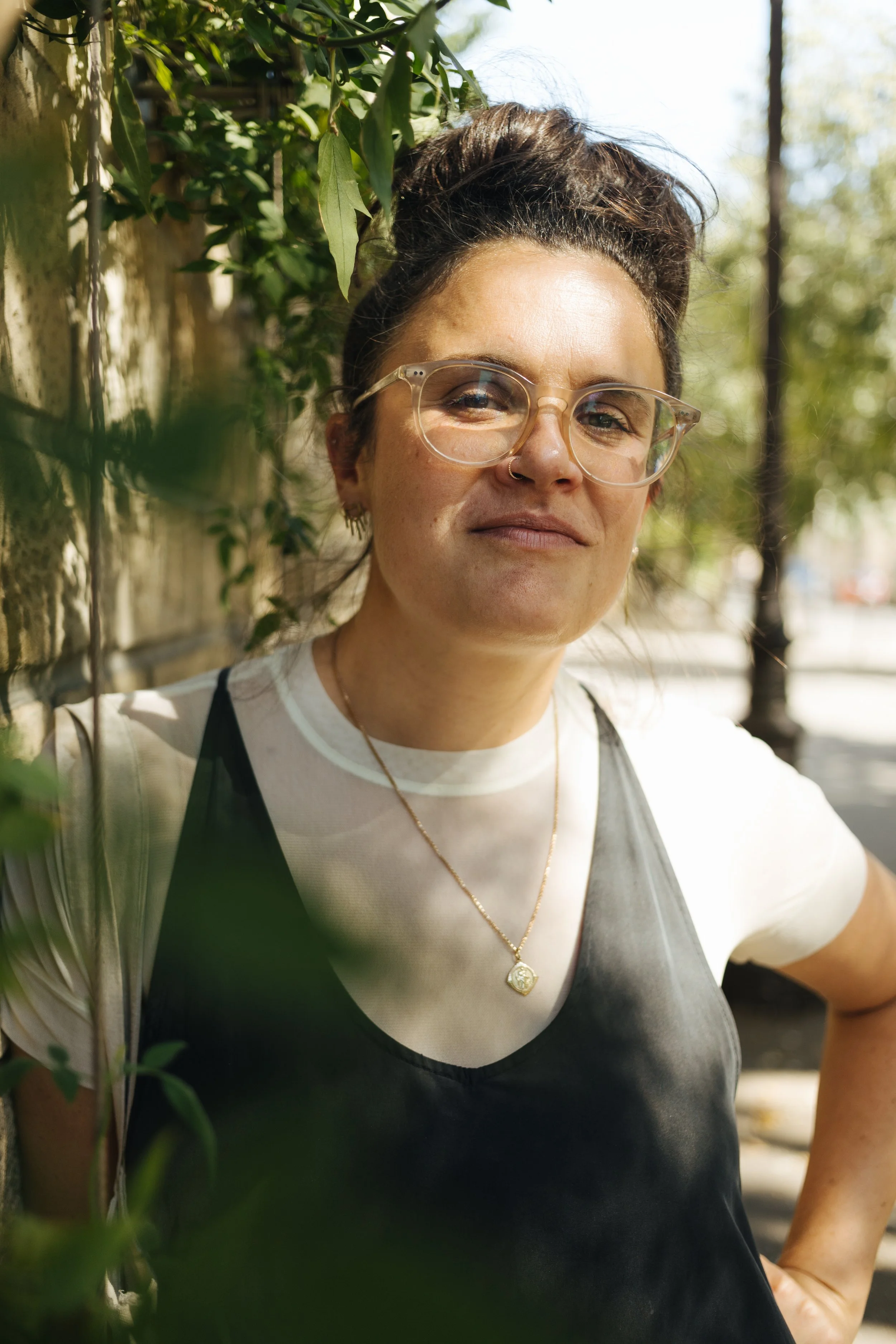 A woman with glasses, hoop earrings, and a gold necklace, standing outdoors near a wall covered with green leaves, sunlight filtering through the trees in the background.