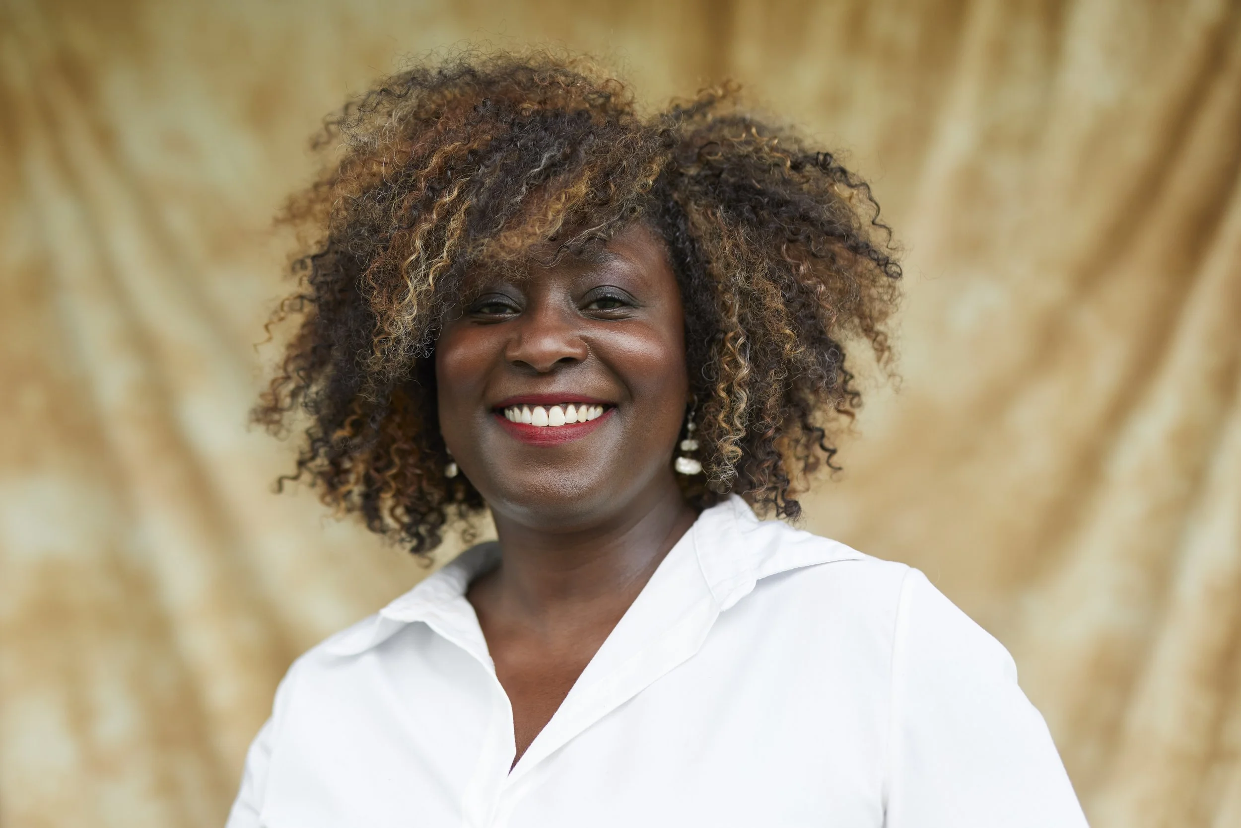 Portrait of a woman with curly hair smiling, wearing a white shirt, with a blurred neutral background.