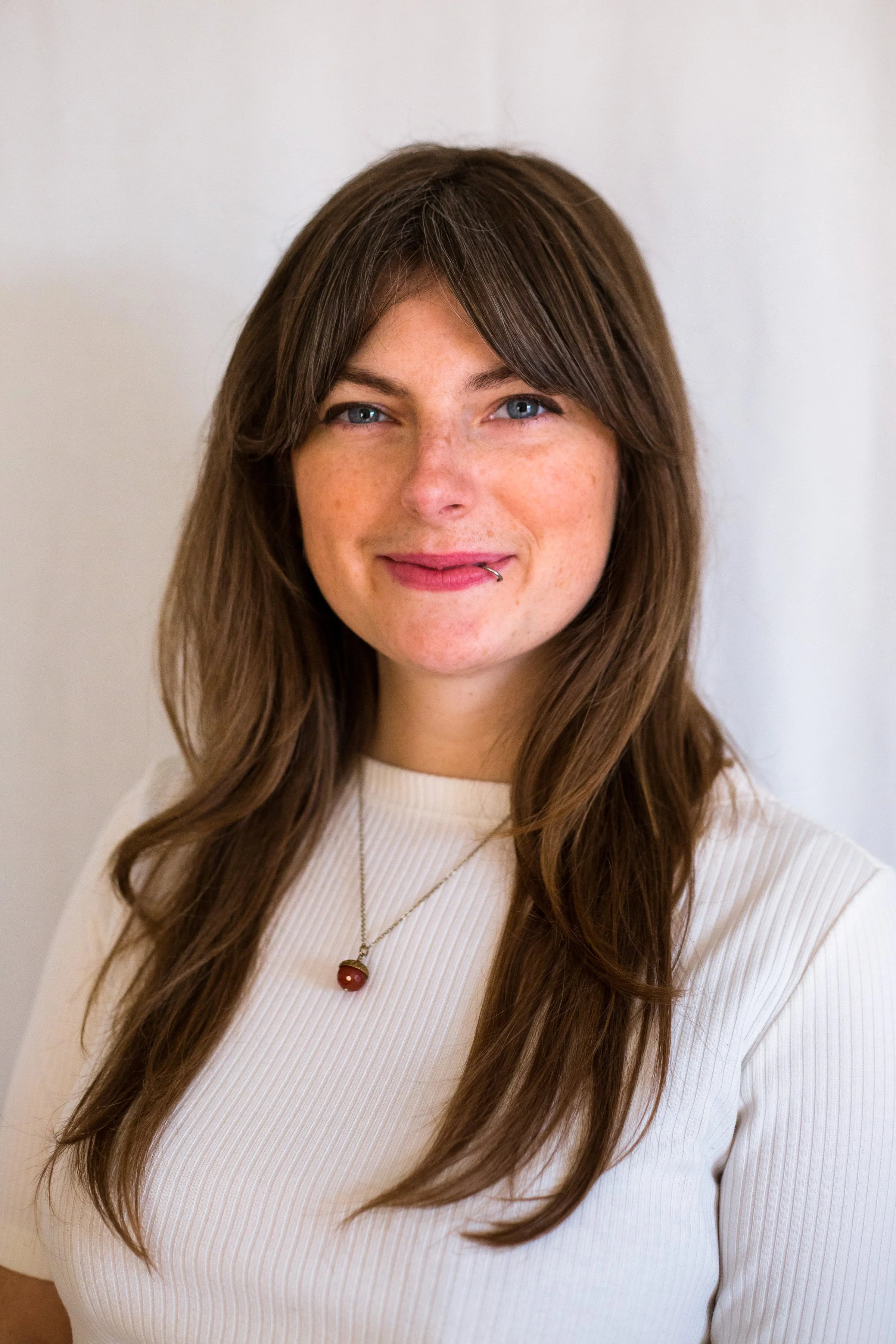 Close-up of a woman with long brown hair, blue eyes, freckles, wearing a white ribbed top, and a necklace with a red gemstone pendant, smiling softly in front of a plain white background.