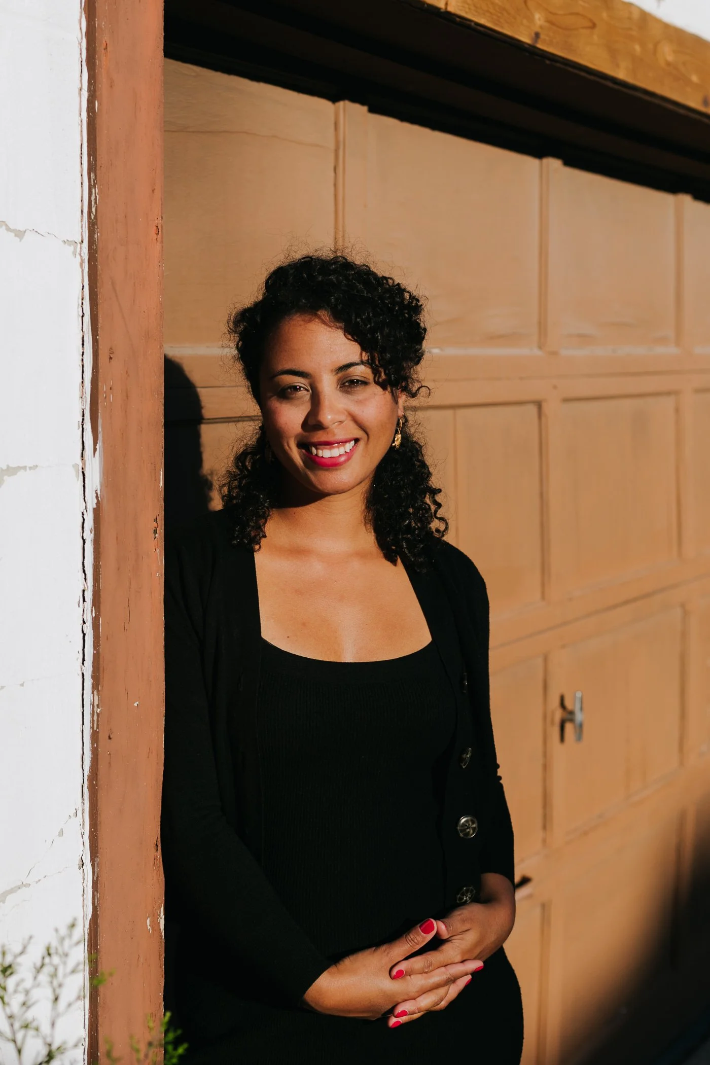 A woman with curly dark hair, wearing earrings and a black top, standing in front of a brown garage door, smiling at the camera.