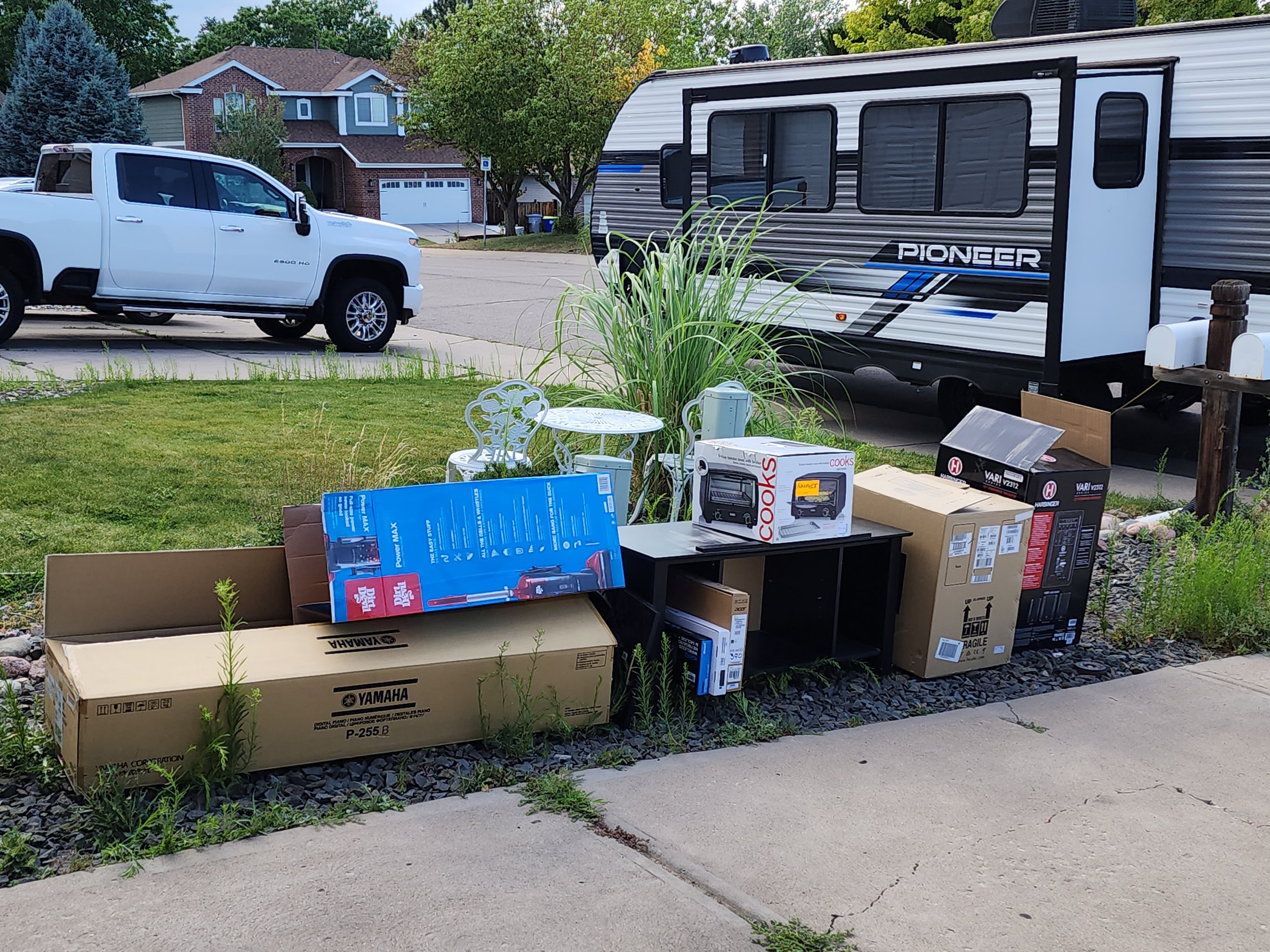 A residential driveway with various boxed appliances and a white pickup truck parked next to a travel trailer, with a house and trees in the background.