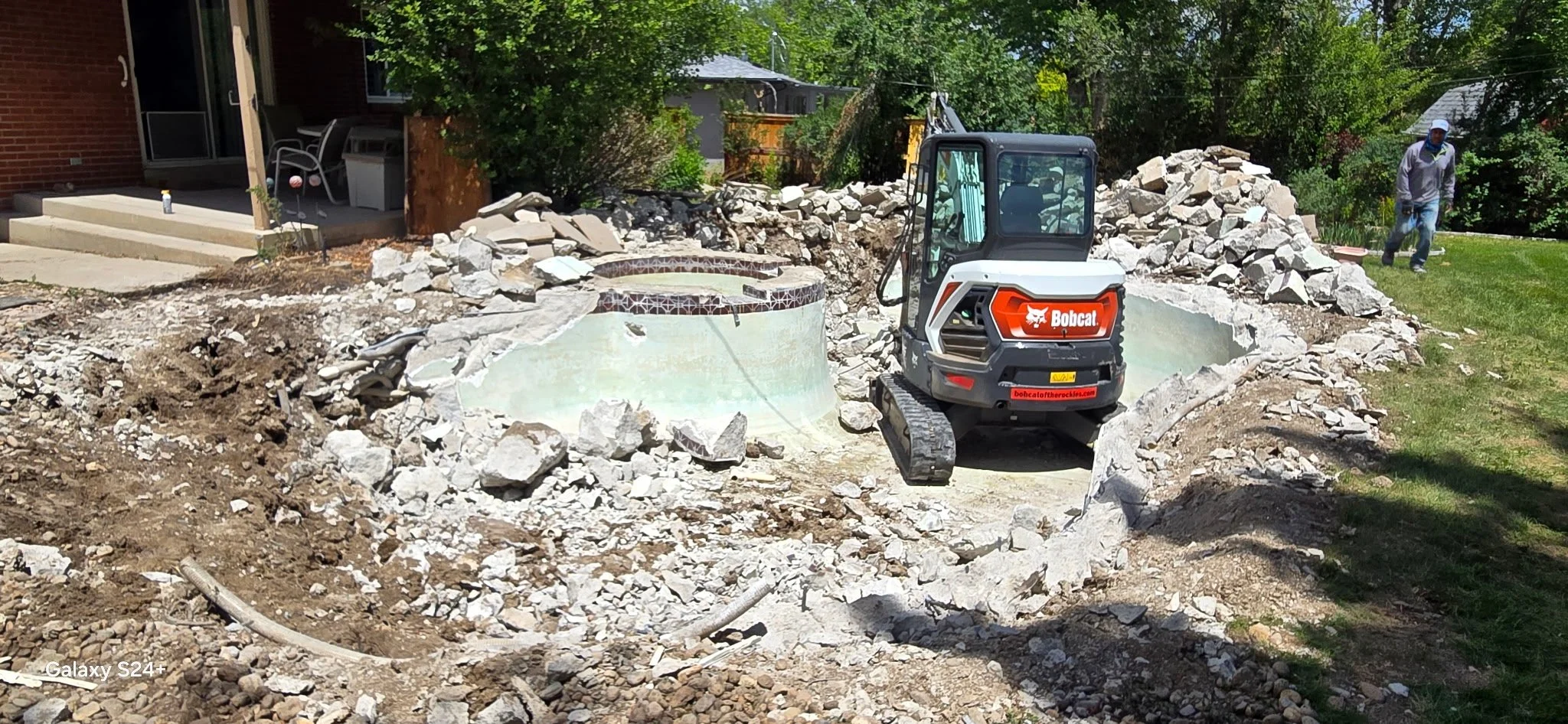 Construction site in a backyard with a Bobcat mini excavator moving rocks and debris, a man walking on the grass, and a patio with chairs and a table near the house.