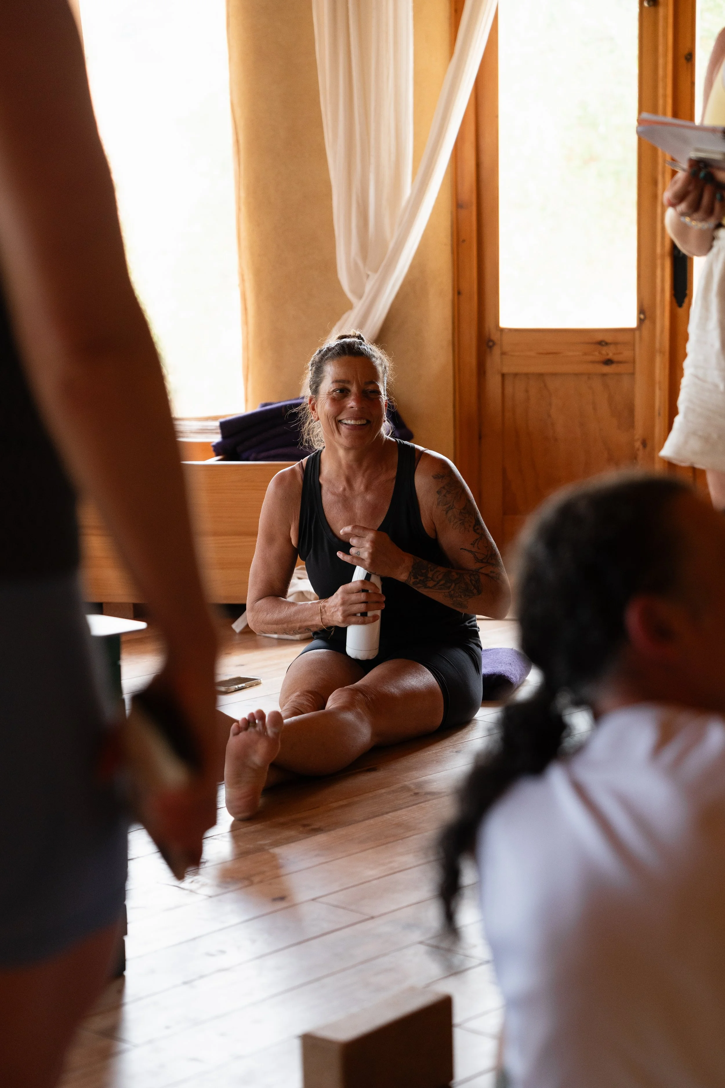 A woman with gray hair tied back, wearing a black tank top, sitting on the wooden floor, smiling and holding a white water bottle, surrounded by young children in a cozy room with wooden walls and large windows.