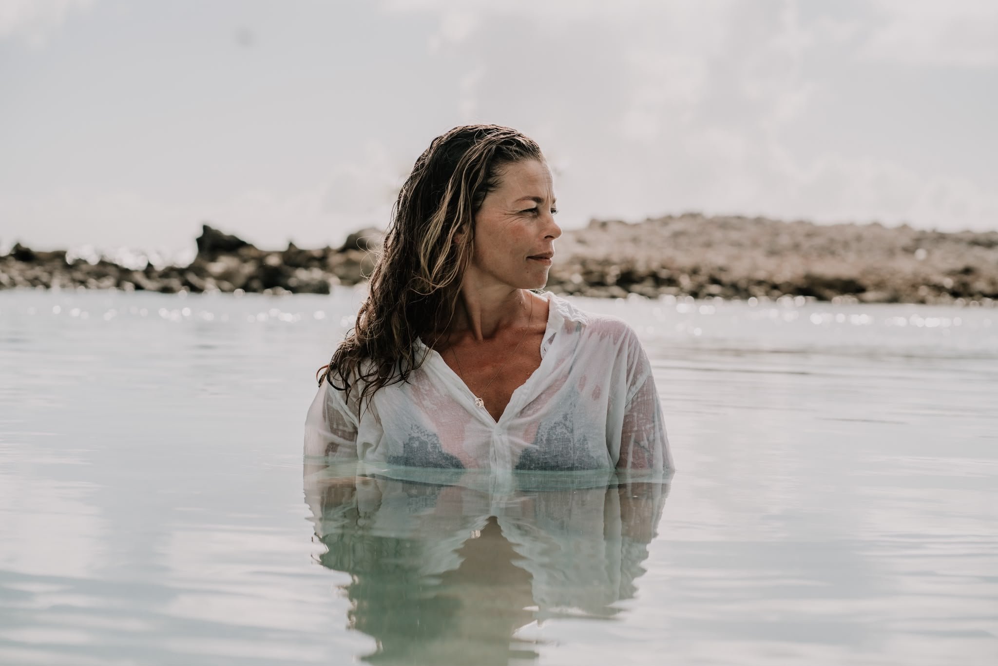 A woman with wet hair standing in shallow water at the beach, looking to the side with rocks and cloudy sky in the background.