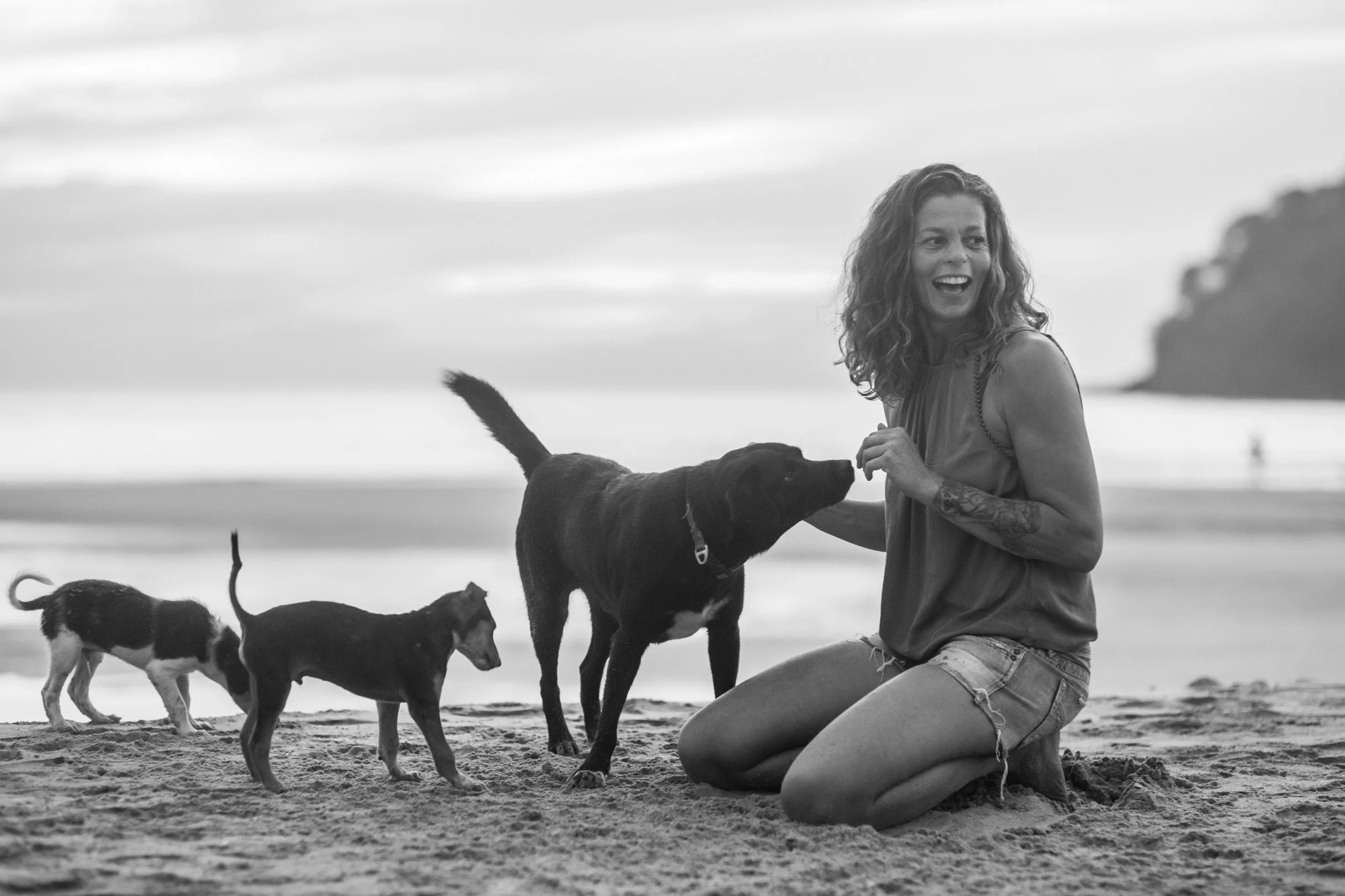 A woman with curly hair and tattoos on her arm, smiling and playing with dogs on a beach at sunset.