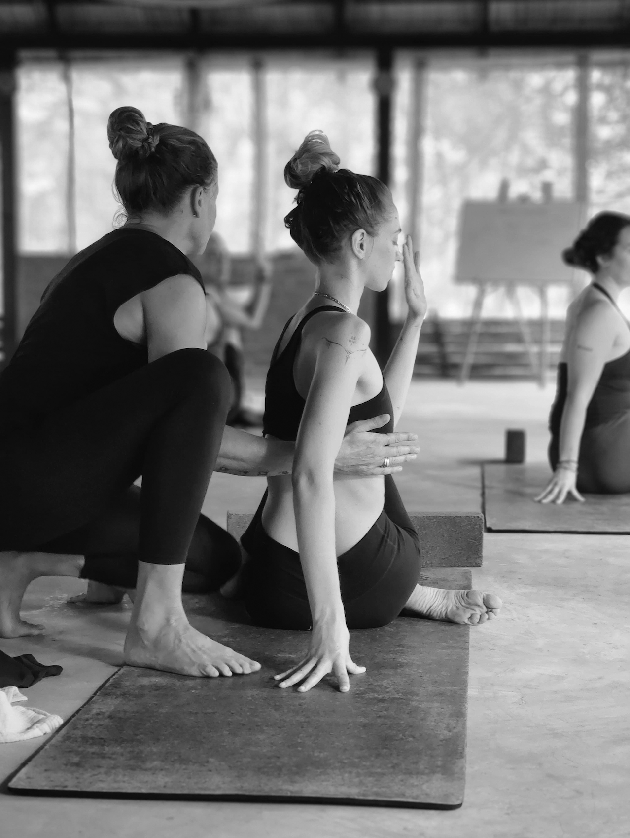 Black and white photo of a woman practicing yoga on a mat, with an instructor assisting her in a lunge pose, in a studio with large windows.