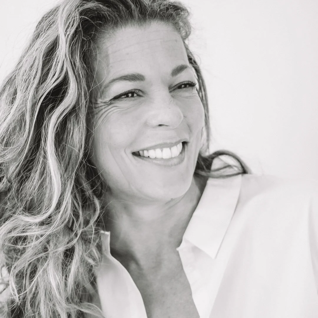 Black and white close-up of a woman smiling with wavy hair, wearing a white collared shirt.