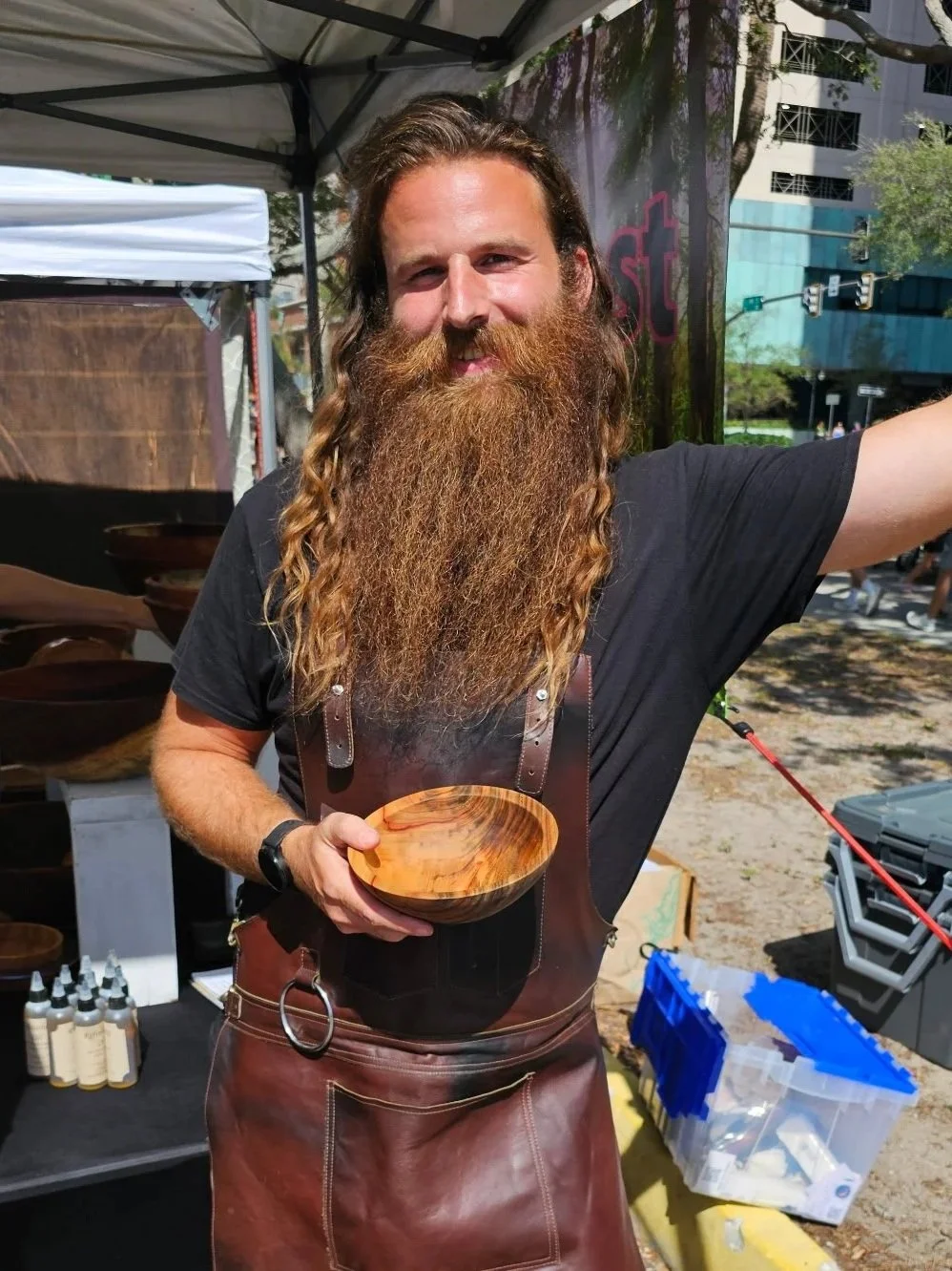 Daniel Marion. Man with long, curly beard and hair, wearing a black t-shirt and brown leather apron, holding a wooden bowl, at an outdoor market or craft fair.