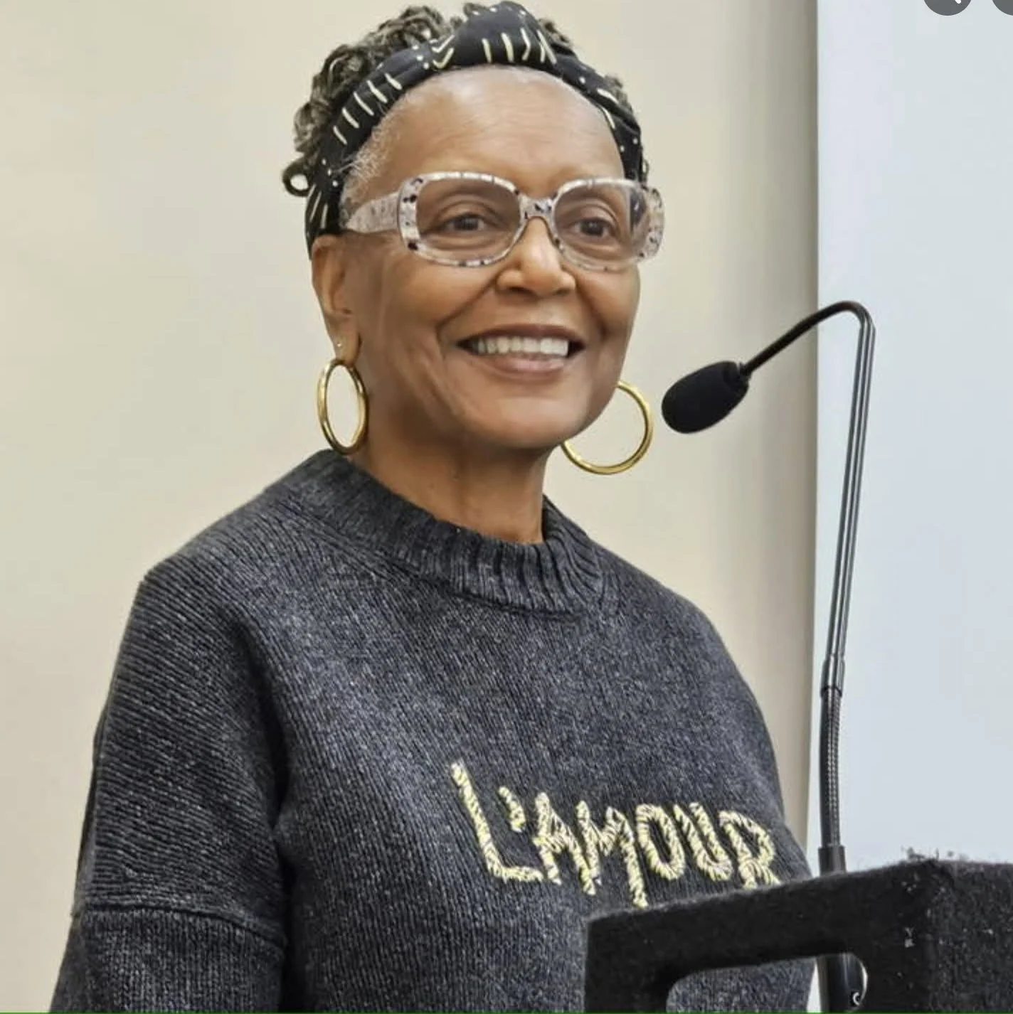 A smiling woman with gray hair, wearing glasses, large hoop earrings, a black patterned headband, and a dark gray sweatshirt with gold lettering, standing in front of a microphone.