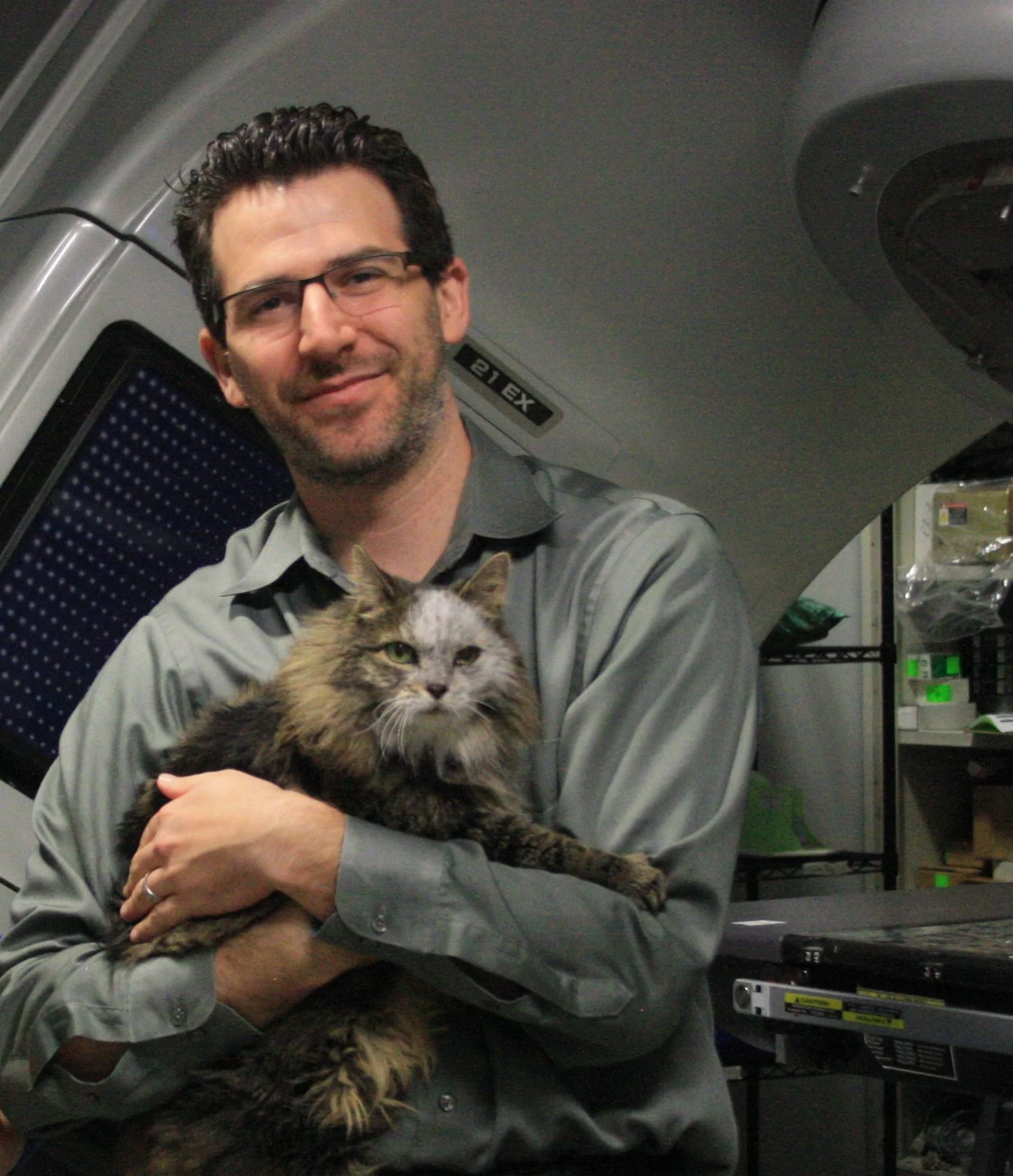 A man with glasses, dark curly hair, and a beard holding a large, long-haired tabby cat inside a room or office with electronic equipment and a gray ceiling.